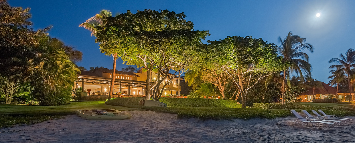 Wide night panorama from the beach: three large floodlit canopy trees dominate the centre, villa glowing behind, full moon top right, beach loungers on the sand, a kayak resting nearby, Casa Manzanillas, Ranchos Estates, Punta Mita.