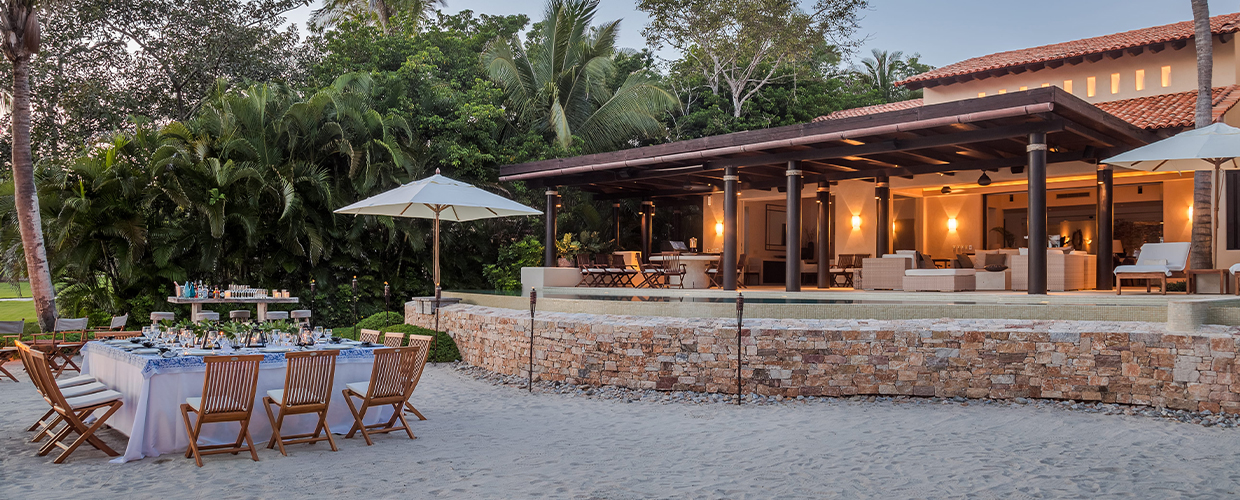 Dusk exterior: a dressed dining table on the sand at left, stone retaining wall, and the villa's open colonnade pavilion warmly lit behind palm-lined grounds, soft amber sky above, Casa Manzanillas, Ranchos Estates, Punta Mita.