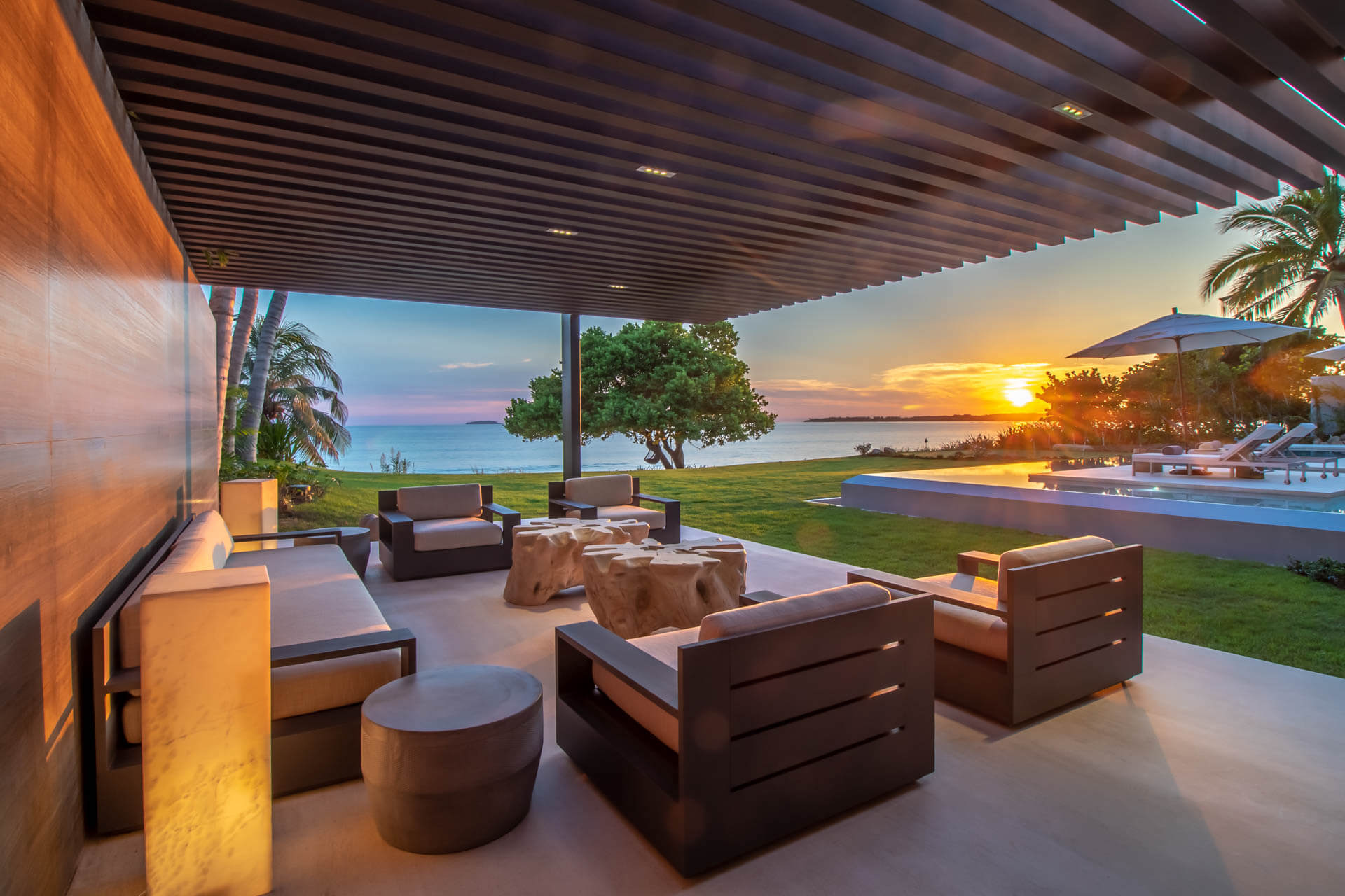 Open-air living pavilion at sunset with dark wood armchairs, driftwood table, pool and ocean beyond, Estate Afrique, Ranchos Estates, Punta Mita.
