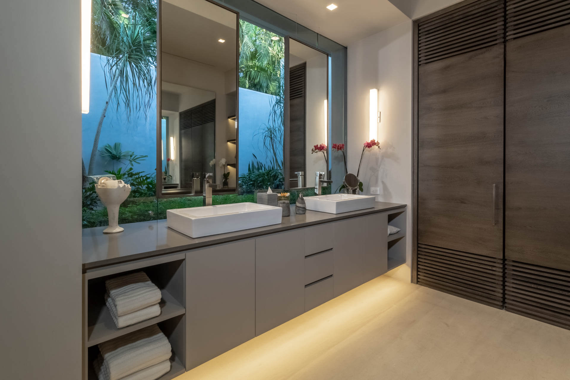 Double-vanity ensuite bath with backlit mirrors, orchid arrangements, and garden window at night, Estate Afrique, Ranchos Estates, Punta Mita.