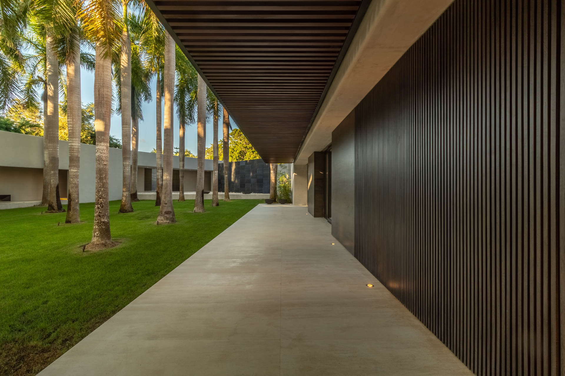 Covered entry corridor with royal palms, cropped lawn, and dark vertical-slat facade at dusk, Estate Afrique, Ranchos Estates, Punta Mita.