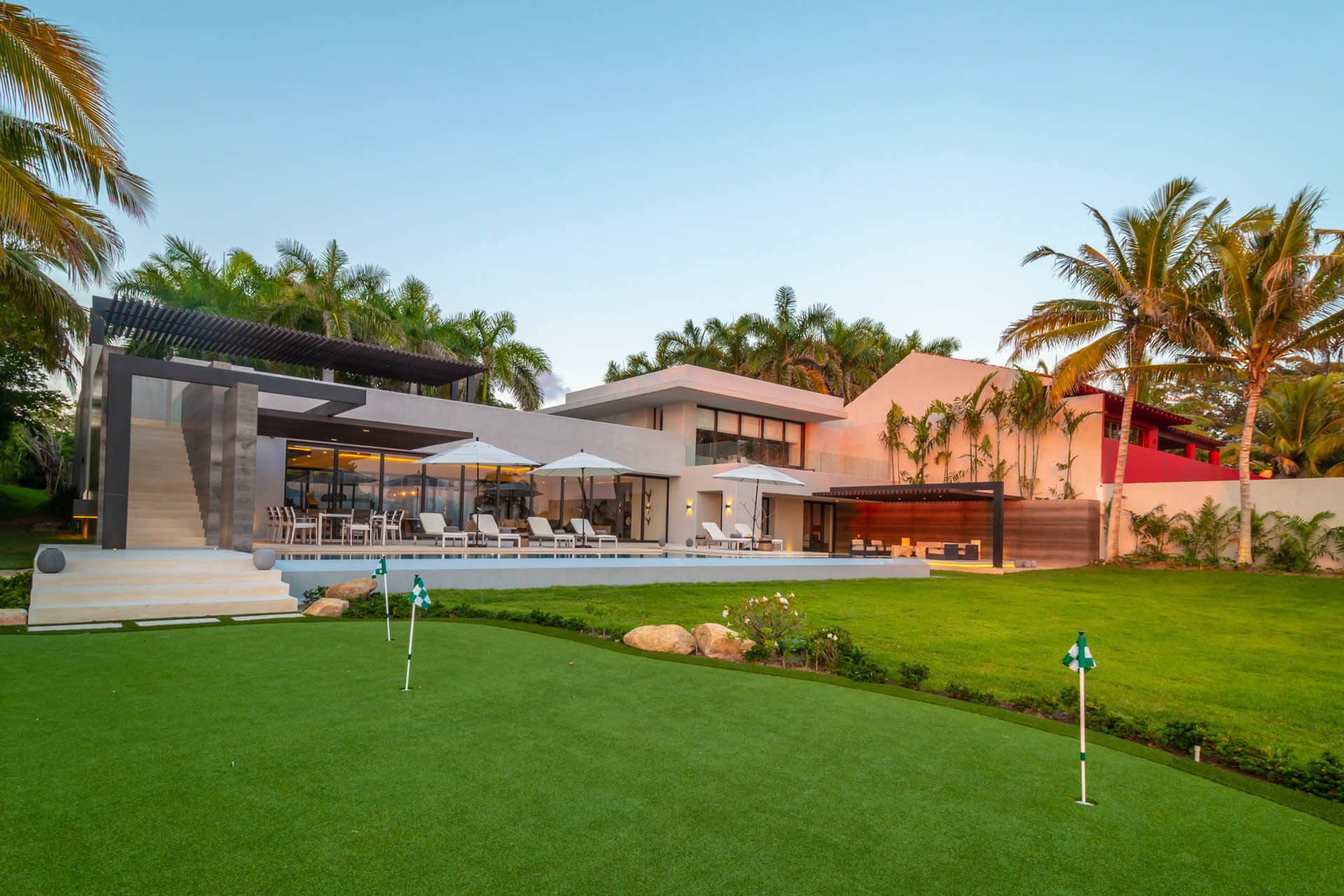 Rear exterior of villa with private putting green, pool terrace with umbrellas, and multi-level concrete facade at dusk, Estate Afrique, Ranchos Estates, Punta Mita.