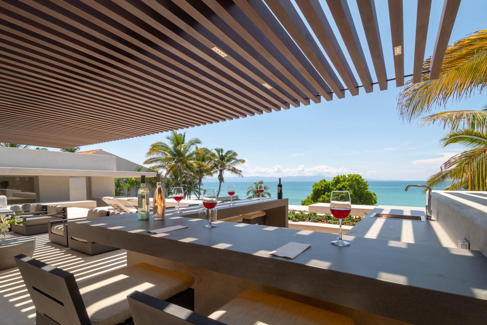 Terrace wet bar counter with two glasses of red wine, wine bottle, and Pacific ocean view under wood-slat pergola, Estate Afrique, Ranchos Estates, Punta Mita.