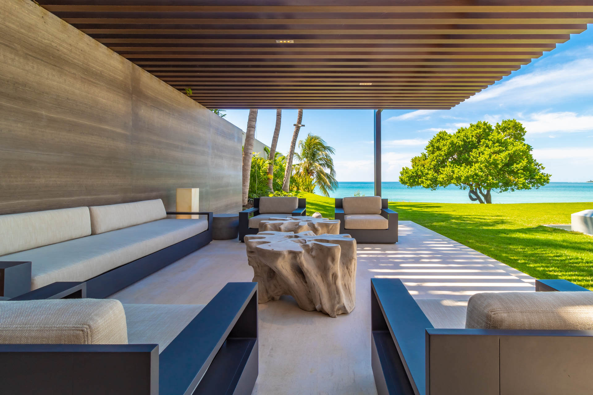 Beachfront living pavilion with driftwood-slab coffee table, linen sofas, and turquoise ocean framed by signature mango tree, Estate Afrique, Ranchos Estates, Punta Mita.