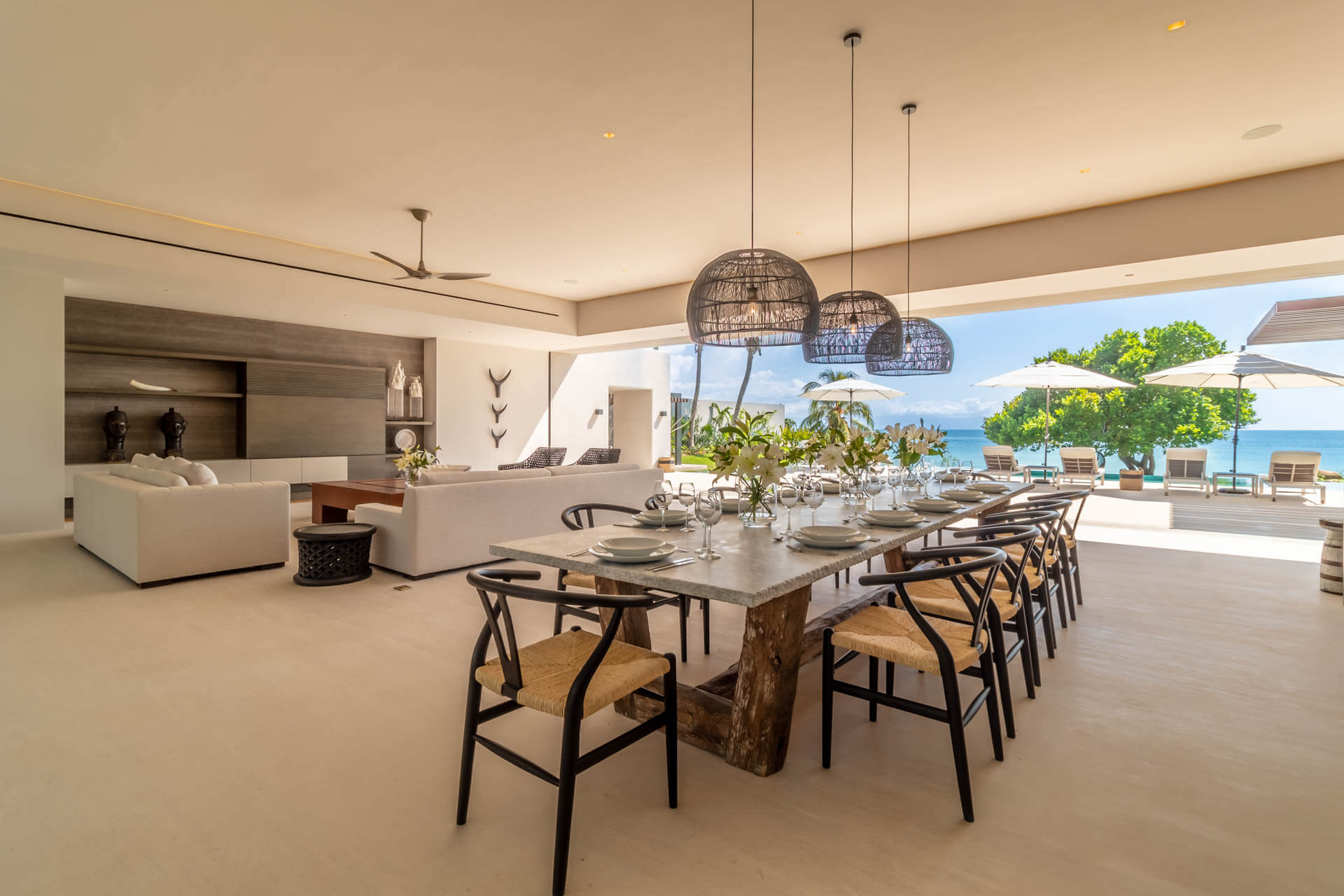 Interior dining room with twelve-seat stone-top table, wishbone chairs, wicker pendant lights, and open wall to pool and Pacific, Estate Afrique, Ranchos Estates, Punta Mita.