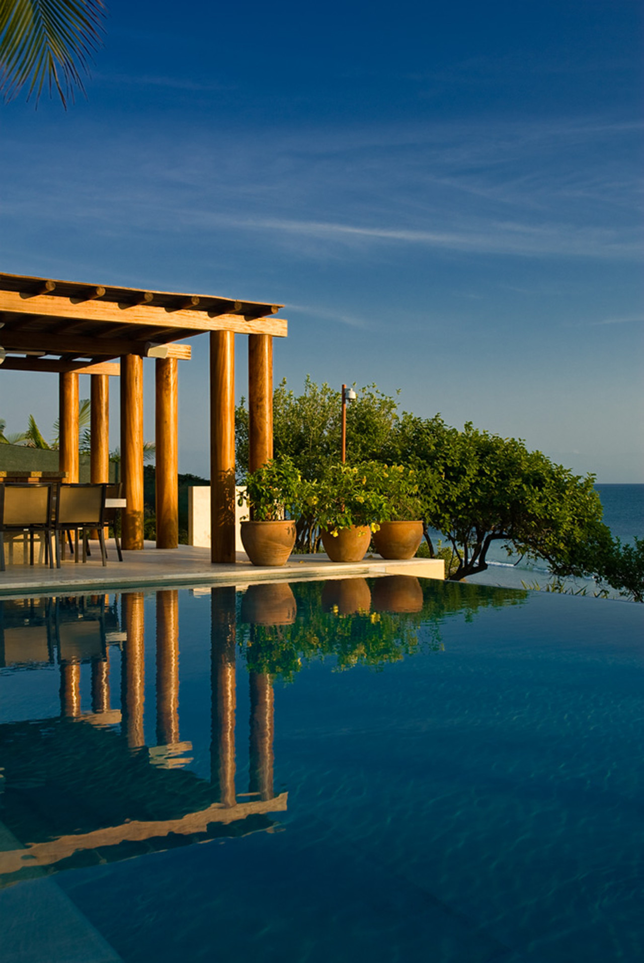 Infinity pool reflecting wood-column palapa with terrace dining chairs and Pacific horizon beyond, Estate Jaguar, Ranchos Estates, Punta Mita.