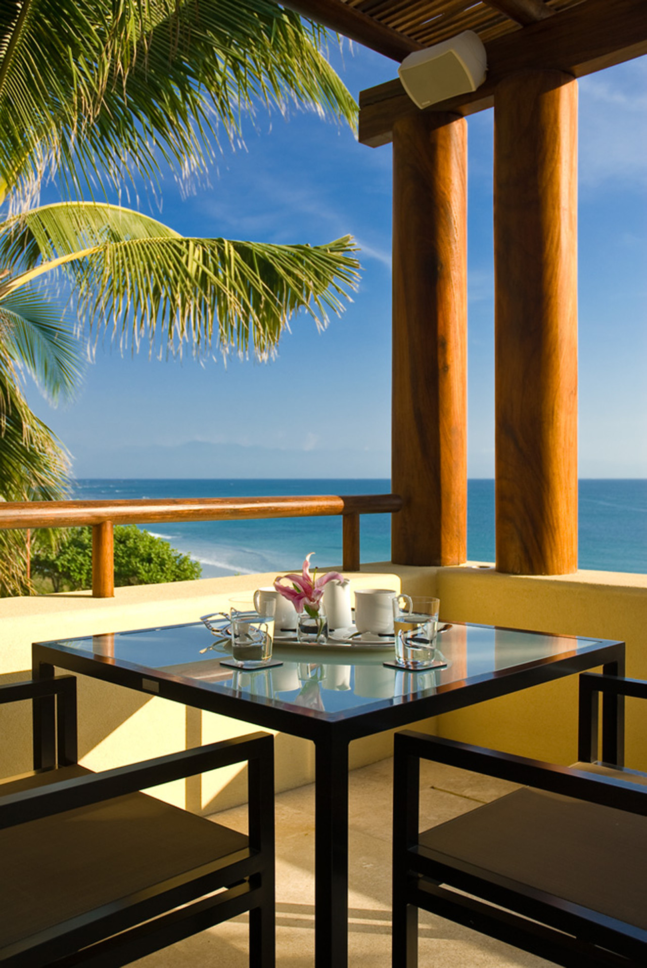Upper terrace breakfast table set for two framed by wood columns and a palm, with ocean and white-sand beach beyond, Estate Jaguar, Ranchos Estates, Punta Mita.