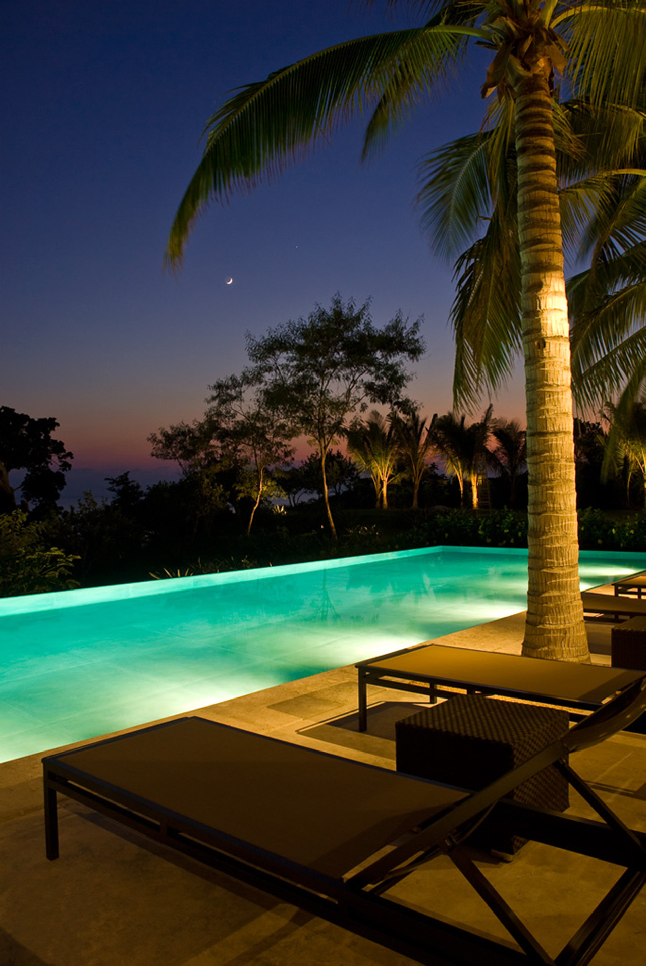 Illuminated infinity pool at dusk with a crescent moon above palm trees and two loungers in the foreground, Estate Jaguar, Ranchos Estates, Punta Mita.