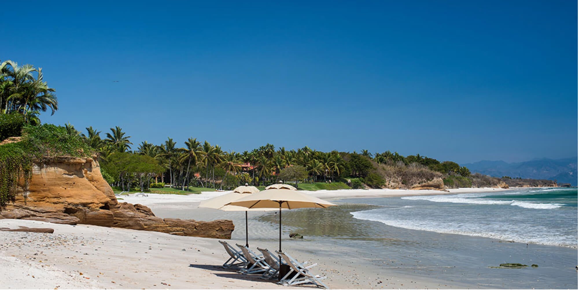 Private beach cove with umbrella and wooden loungers beside red sandstone cliffs, villa visible in the palm treeline above, Estate Jaguar, Ranchos Estates, Punta Mita.