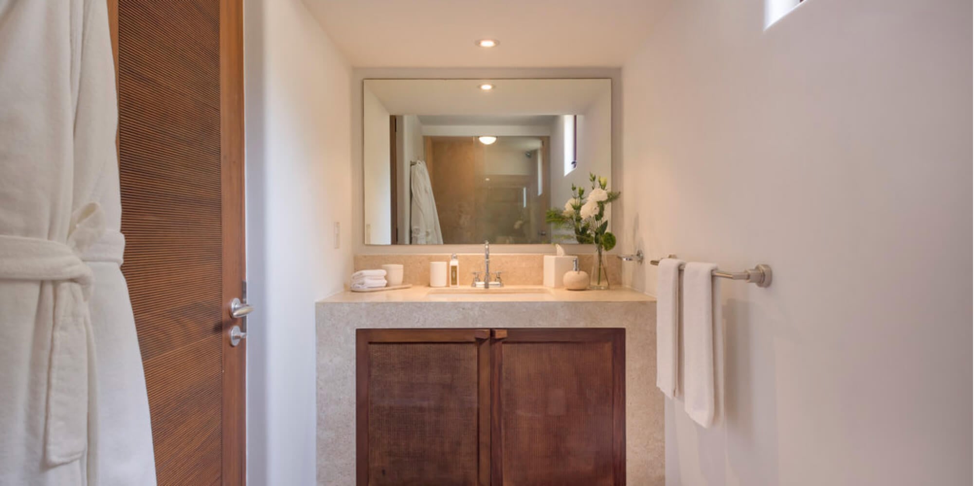 Guest ensuite bathroom with travertine vanity, carved wood cabinet below, framed mirror, folded towels, and a vase of white roses, Estate Jaguar, Ranchos Estates, Punta Mita.