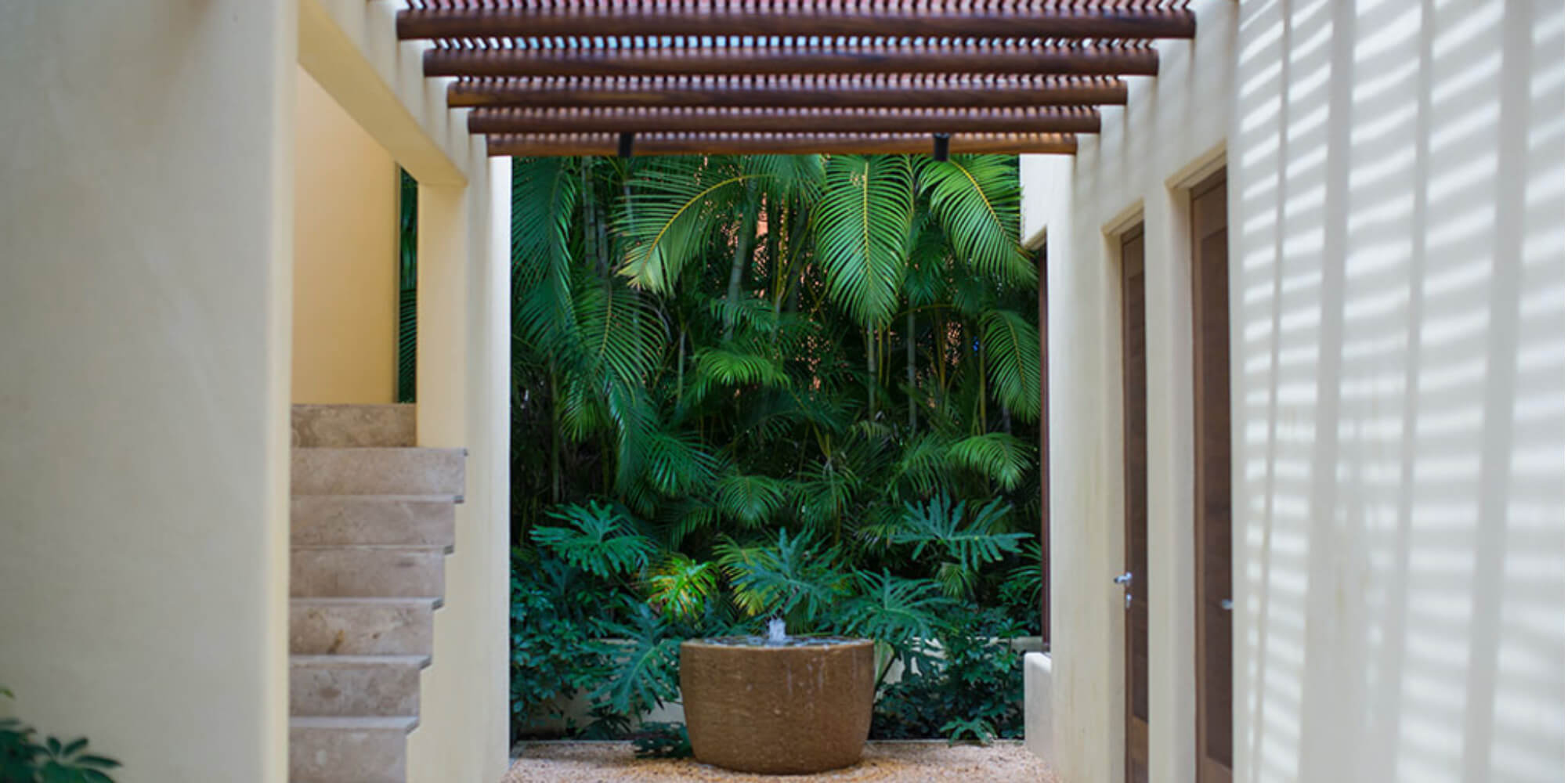 Interior courtyard corridor with travertine stairs, cream stucco walls with slatted shutters, and a dense tropical palm garden framed in the opening, Estate Jaguar, Ranchos Estates, Punta Mita.