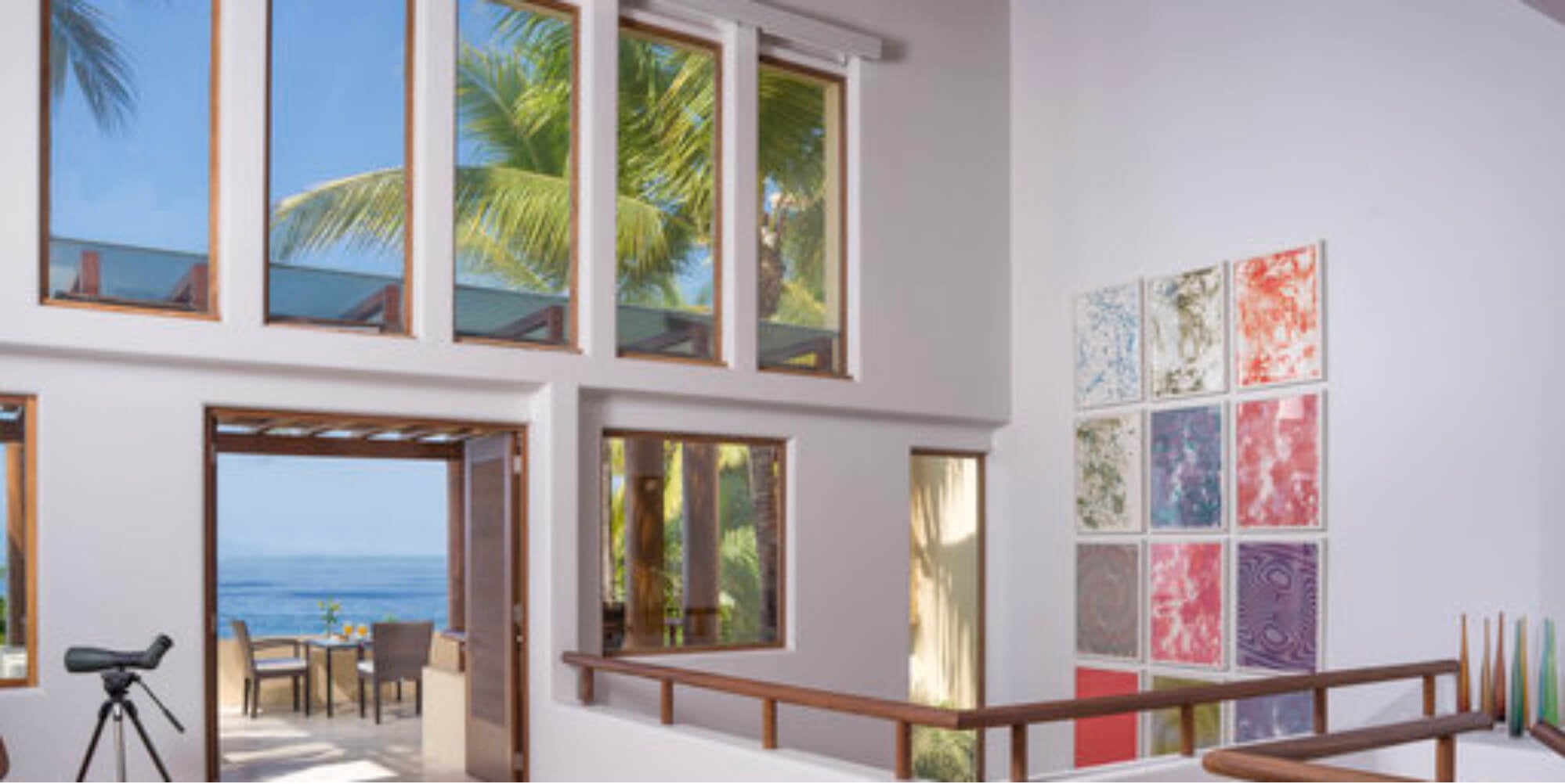 Upper landing of the double-height living pavilion with the twelve-panel art grid, open terrace doors framing the ocean, and a spotting scope on a tripod, Estate Jaguar, Ranchos Estates, Punta Mita.