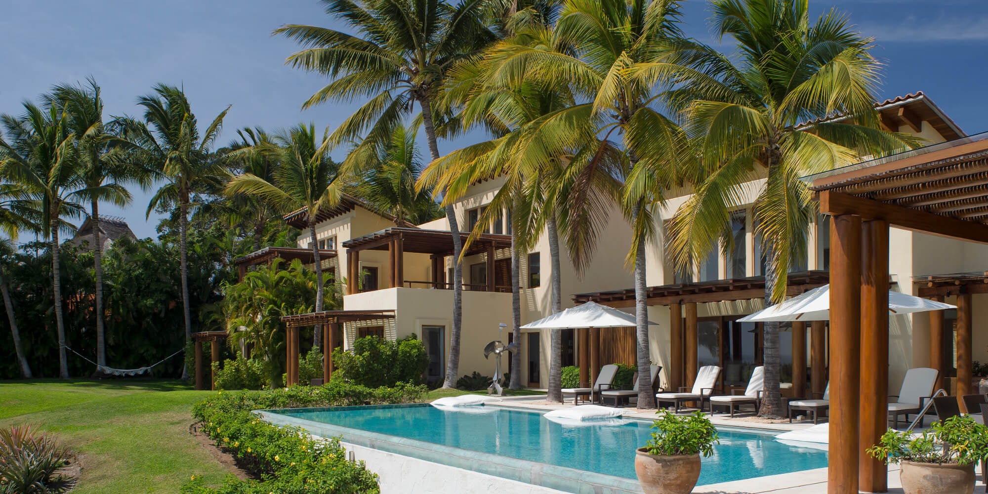 Pool terrace and main building facade with loungers under umbrella, wood-column pergola, and coconut palms against a blue sky, Estate Jaguar, Ranchos Estates, Punta Mita.