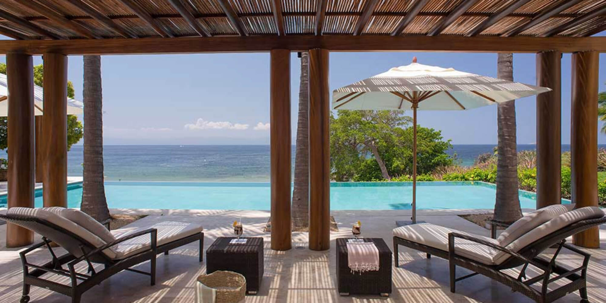 Shaded palapa lounge with two rattan chaise longues under a bamboo-slat ceiling, looking through wood columns to the infinity pool and Pacific, Estate Jaguar, Ranchos Estates, Punta Mita.
