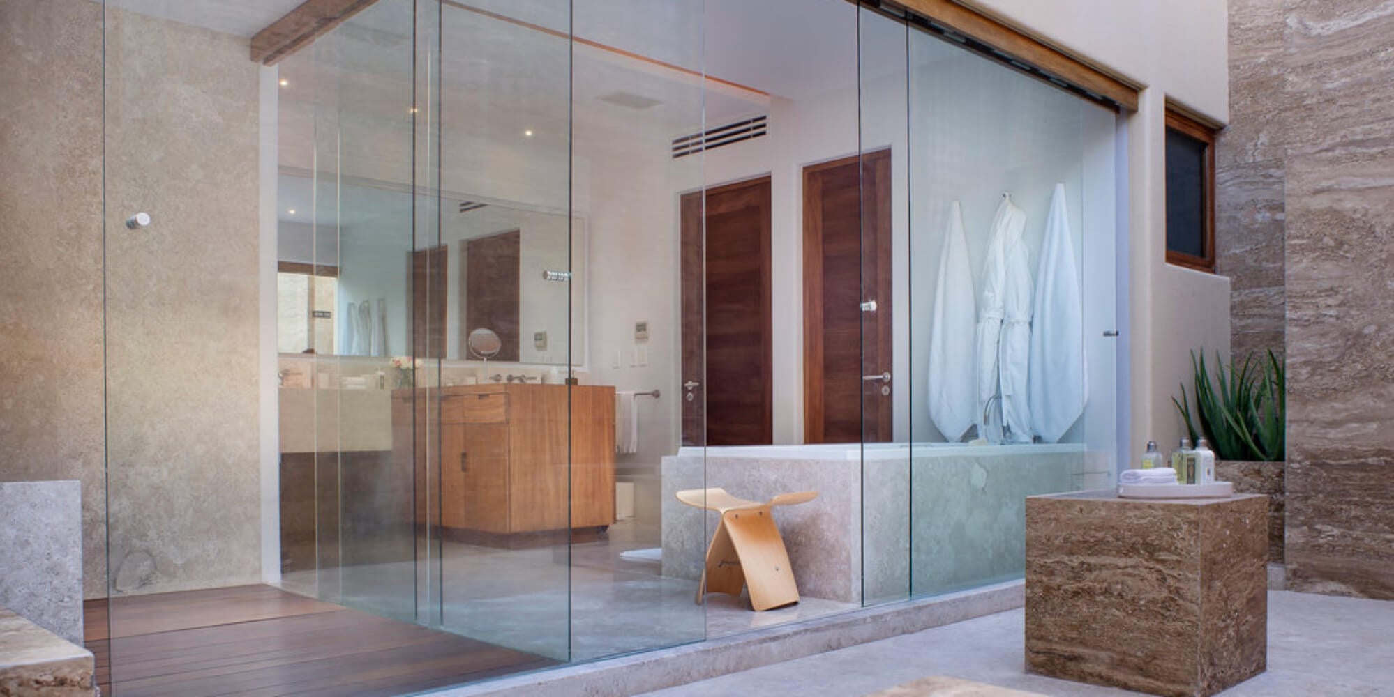 Primary suite spa bathroom with a full glass wall enclosure, travertine soaking tub, round wood vanity, two robes, and an outdoor aloe planter, Estate Jaguar, Ranchos Estates, Punta Mita.