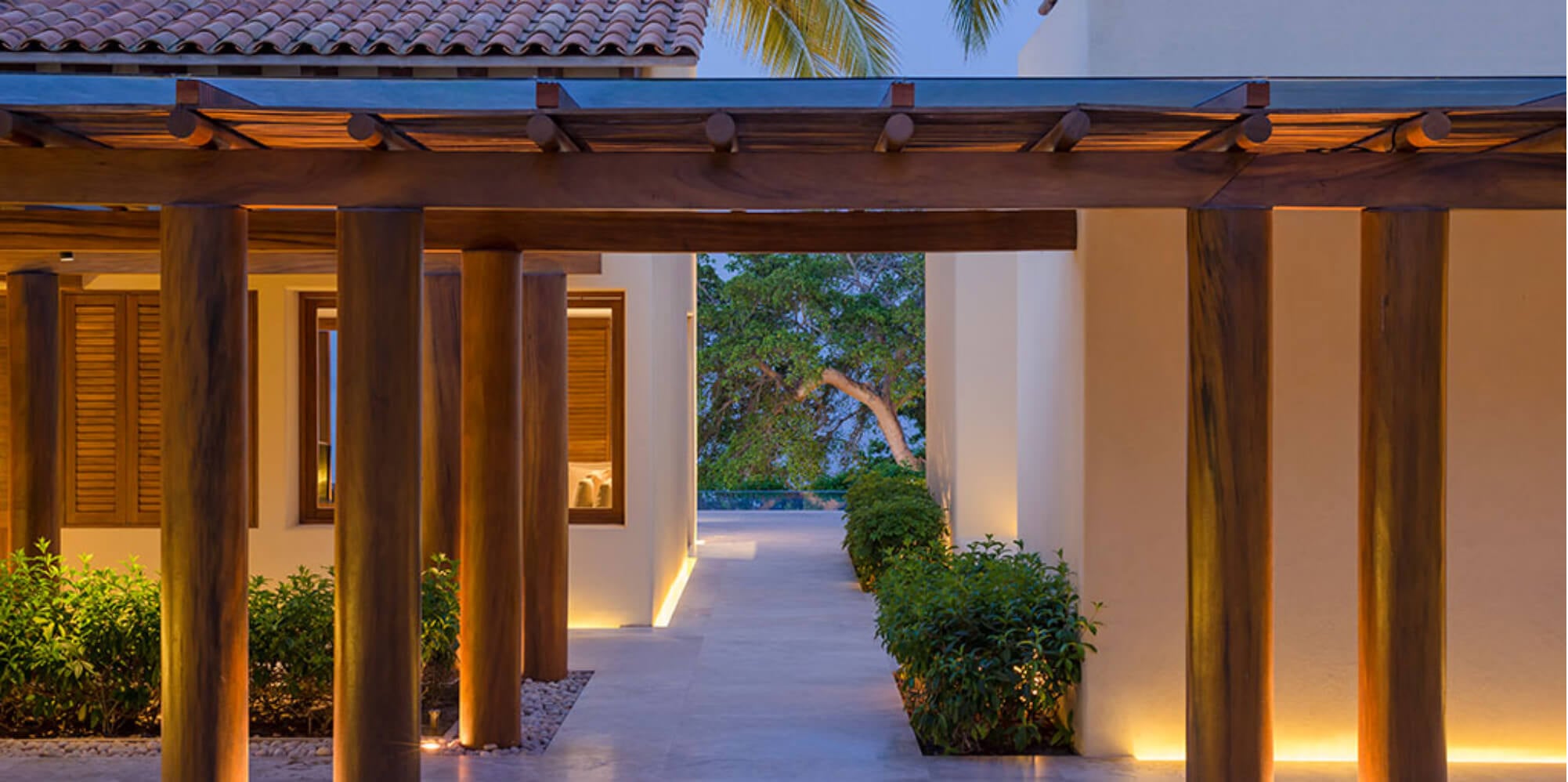 Covered entry walkway with warm uplighting on wood pergola columns and cream stucco walls at dusk, Estate Jaguar, Ranchos Estates, Punta Mita.