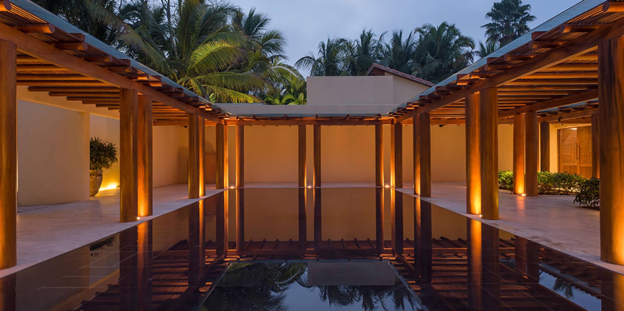 Open-air pavilion with a dark reflecting pool flanked by wood columns glowing with amber uplighting at twilight, Estate Jaguar, Ranchos Estates, Punta Mita.