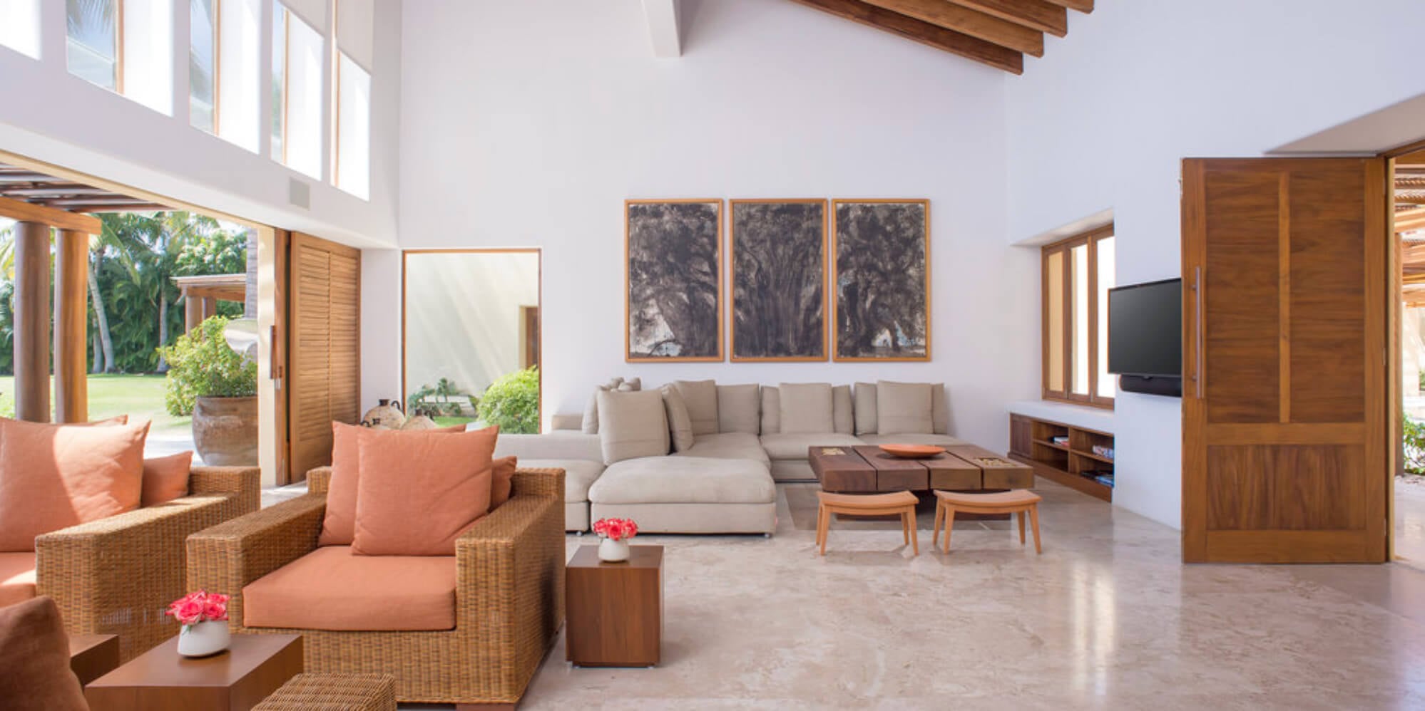 High-ceilinged living room with exposed wood beams, rattan armchairs in coral upholstery, a cream sectional sofa, and louvered doors open to the garden, Estate Jaguar, Ranchos Estates, Punta Mita.