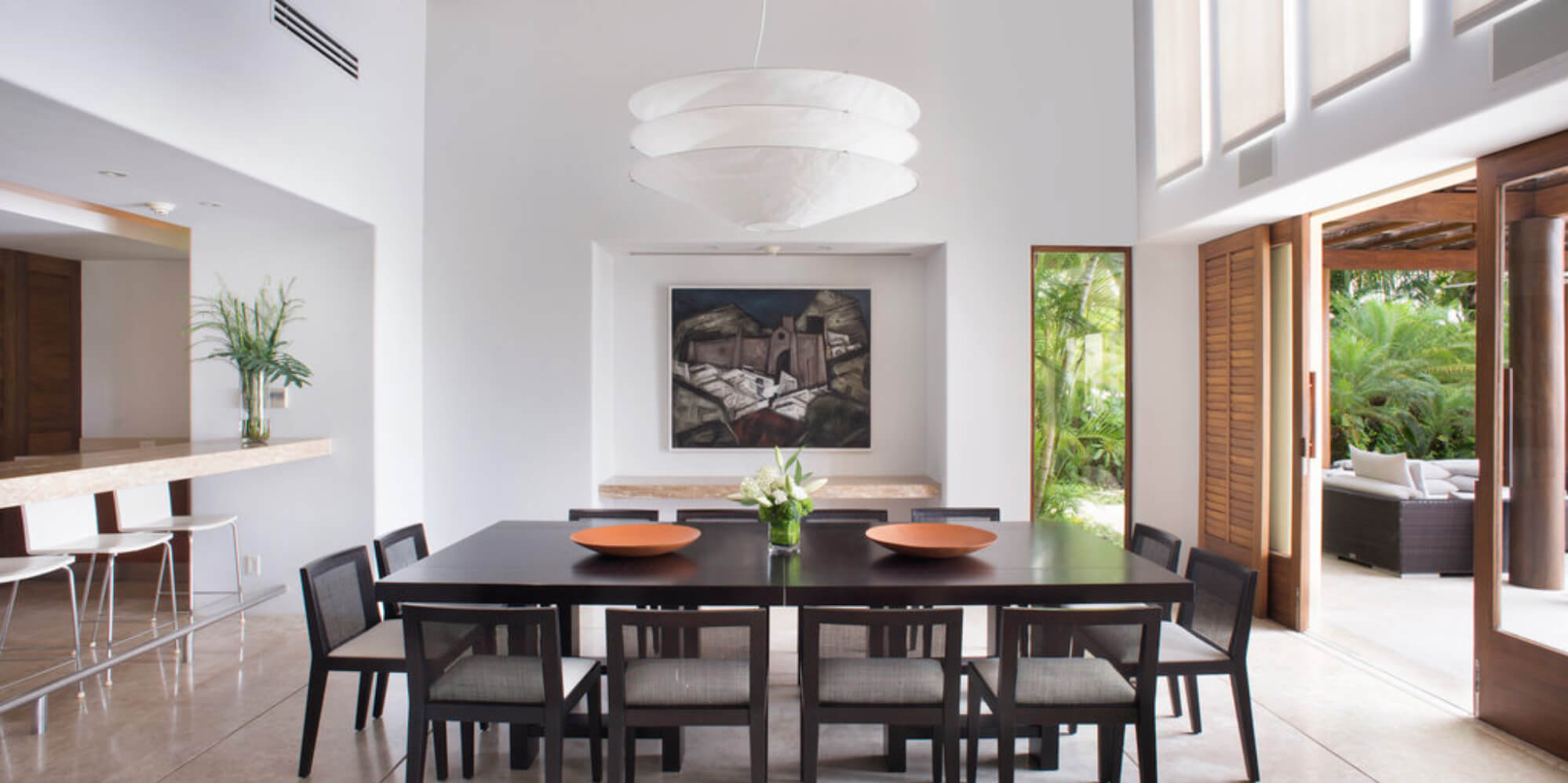 Indoor dining room with a dark wood table set for eight, a large sculptural pendant lamp, a framed painting on the wall, and louvered doors open to the garden, Estate Jaguar, Ranchos Estates, Punta Mita.