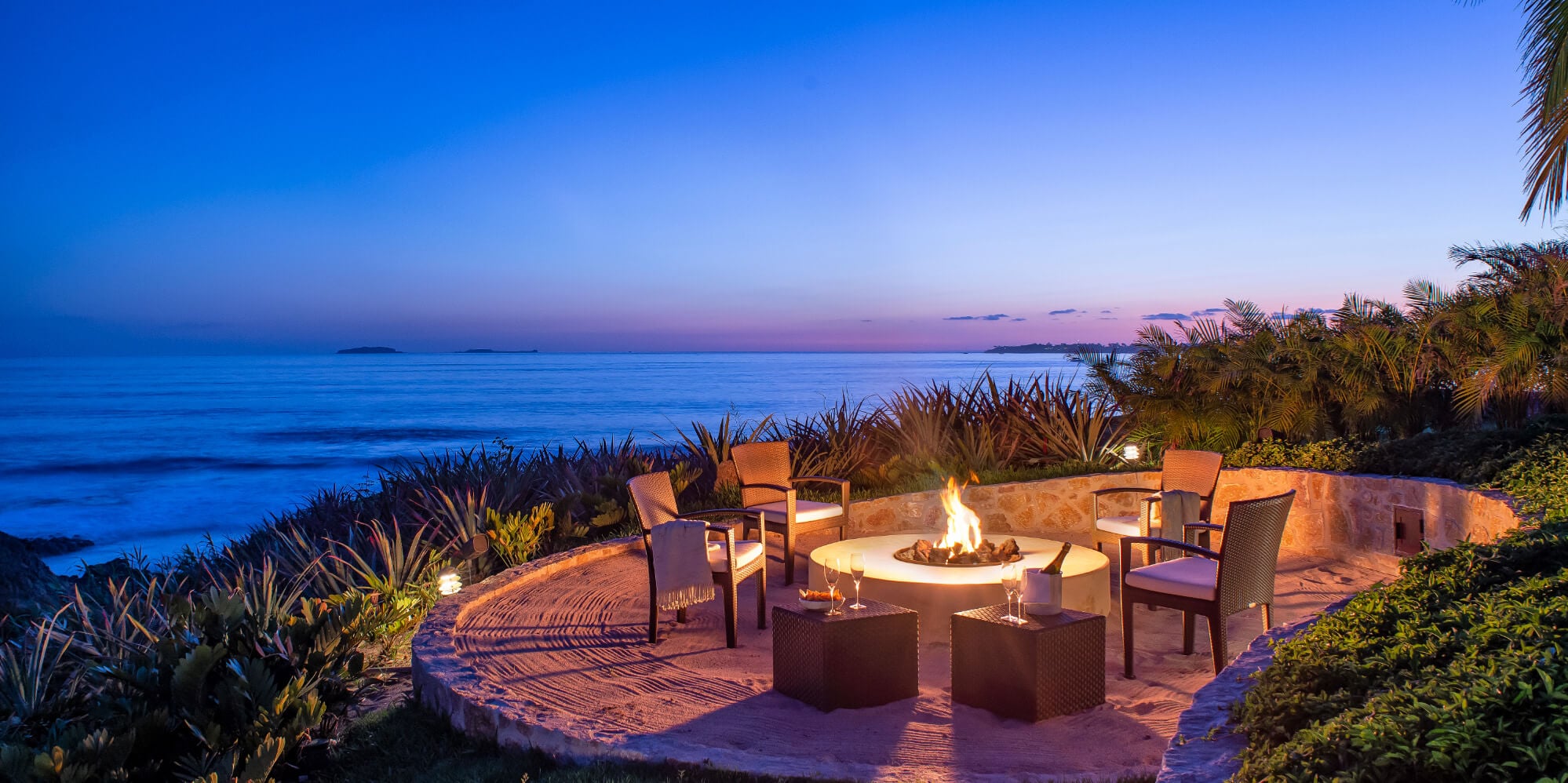 Circular stone fire-pit terrace at dusk with rattan chairs, agave plantings, and the Pacific Ocean glowing blue at the horizon, Estate Jaguar, Ranchos Estates, Punta Mita.
