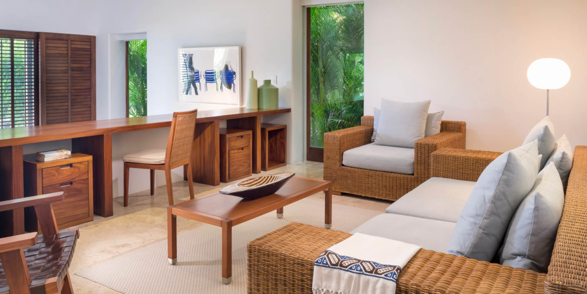 Guest suite sitting room with rattan sofa and armchair, a wood writing desk, colorful artwork, and sliding doors open to a garden terrace, Estate Jaguar, Ranchos Estates, Punta Mita.