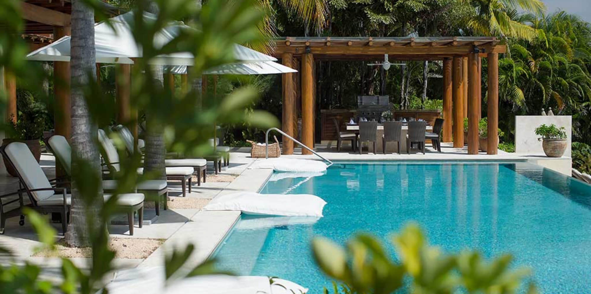 Pool deck with white float mats in the water, sun loungers under umbrellas, and a wood pergola sheltering an outdoor dining table, Estate Jaguar, Ranchos Estates, Punta Mita.