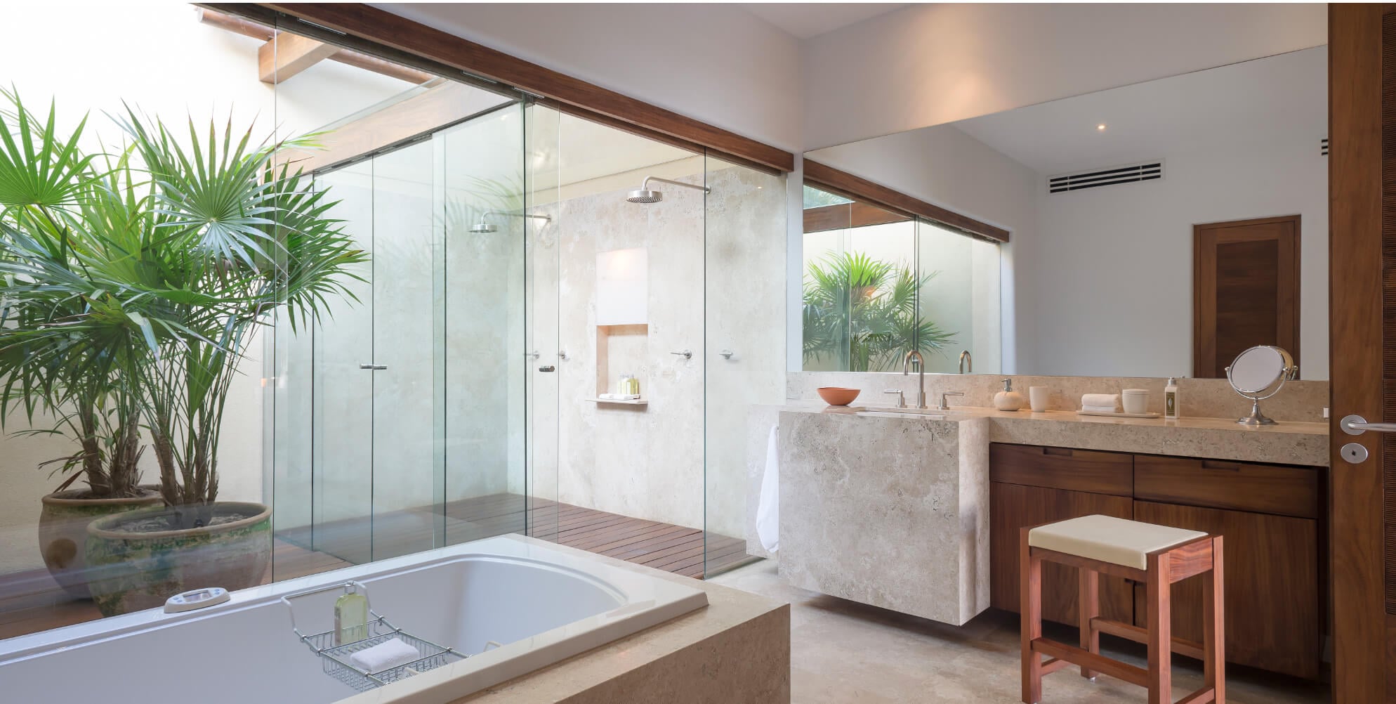 Primary bathroom with a travertine soaking tub, a glass walk-in shower with rain head and wood-slatted mat, a stone double vanity, and a potted palm beside garden windows, Estate Jaguar, Ranchos Estates, Punta Mita.