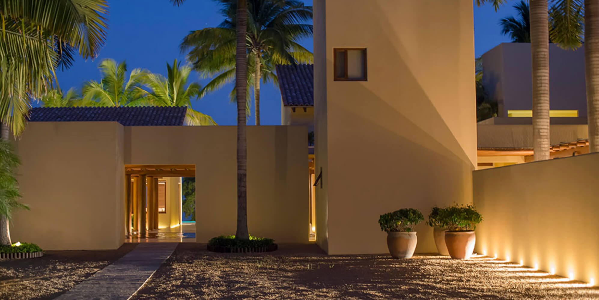 Arrival courtyard at dusk with tall cream stucco walls lit by uplights, terracotta pots with clipped topiaries, and palms against a deep blue sky, Estate Jaguar, Ranchos Estates, Punta Mita.