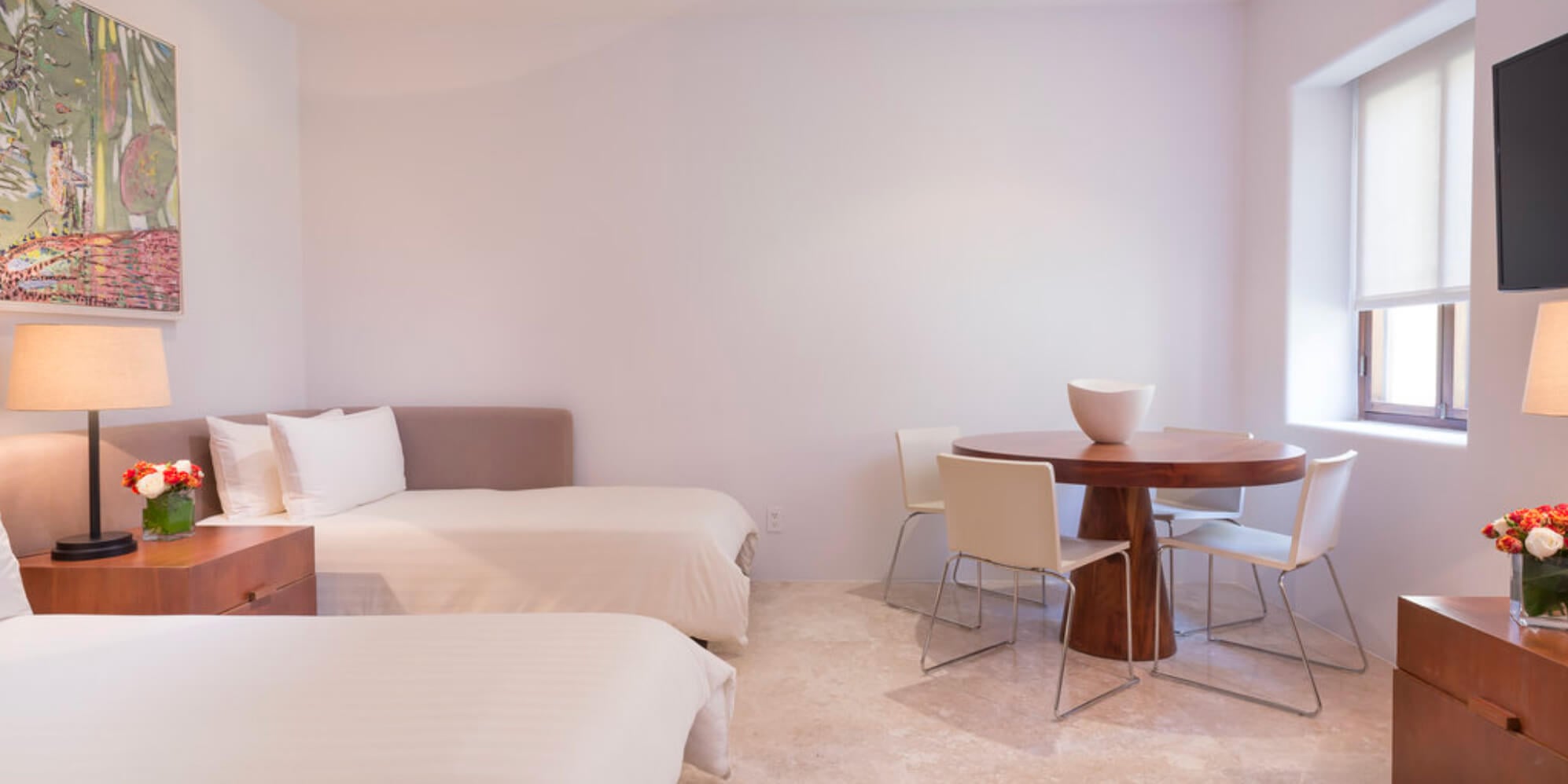 Guest suite with two queen beds, a round wood dining table with four cream chairs, colorful artwork, and a flat-screen television beside a window, Estate Jaguar, Ranchos Estates, Punta Mita.