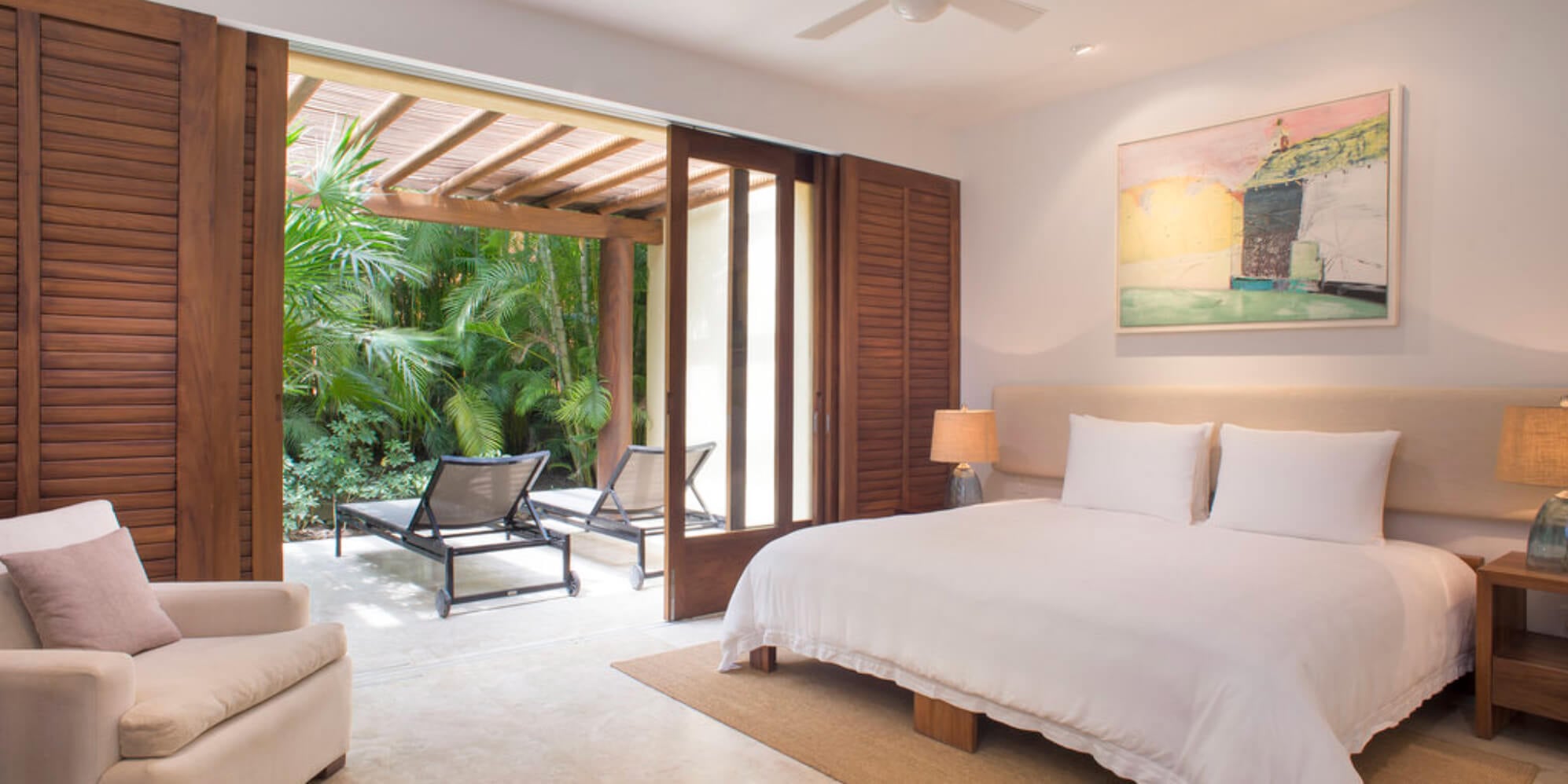 Guest bedroom with a king bed, upholstered headboard, and louvered wood doors open to a private terrace with sun loungers in a jungle garden, Estate Jaguar, Ranchos Estates, Punta Mita.