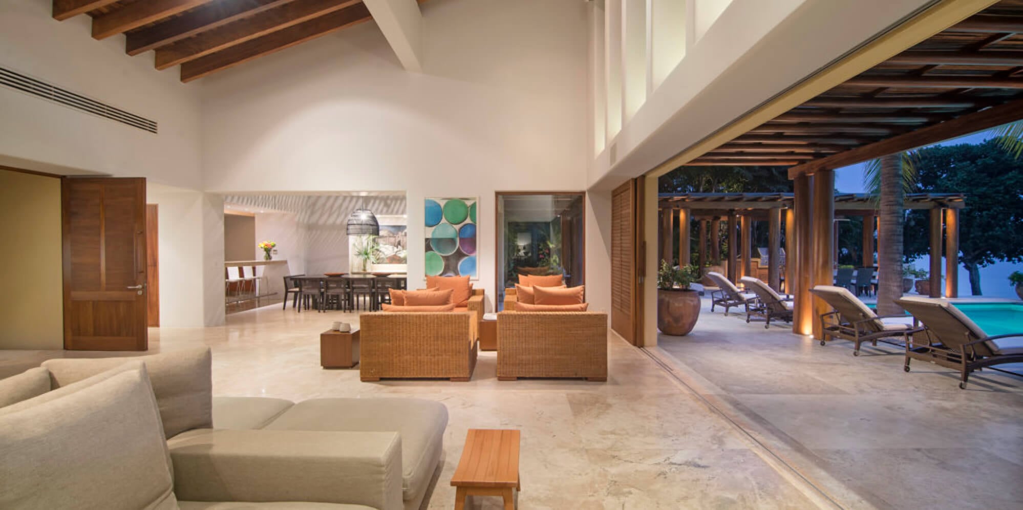 Open-plan living pavilion at dusk showing the living area with rattan sofas, the dining room beyond, and the pool terrace with loungers lit through fully open doors, Estate Jaguar, Ranchos Estates, Punta Mita.