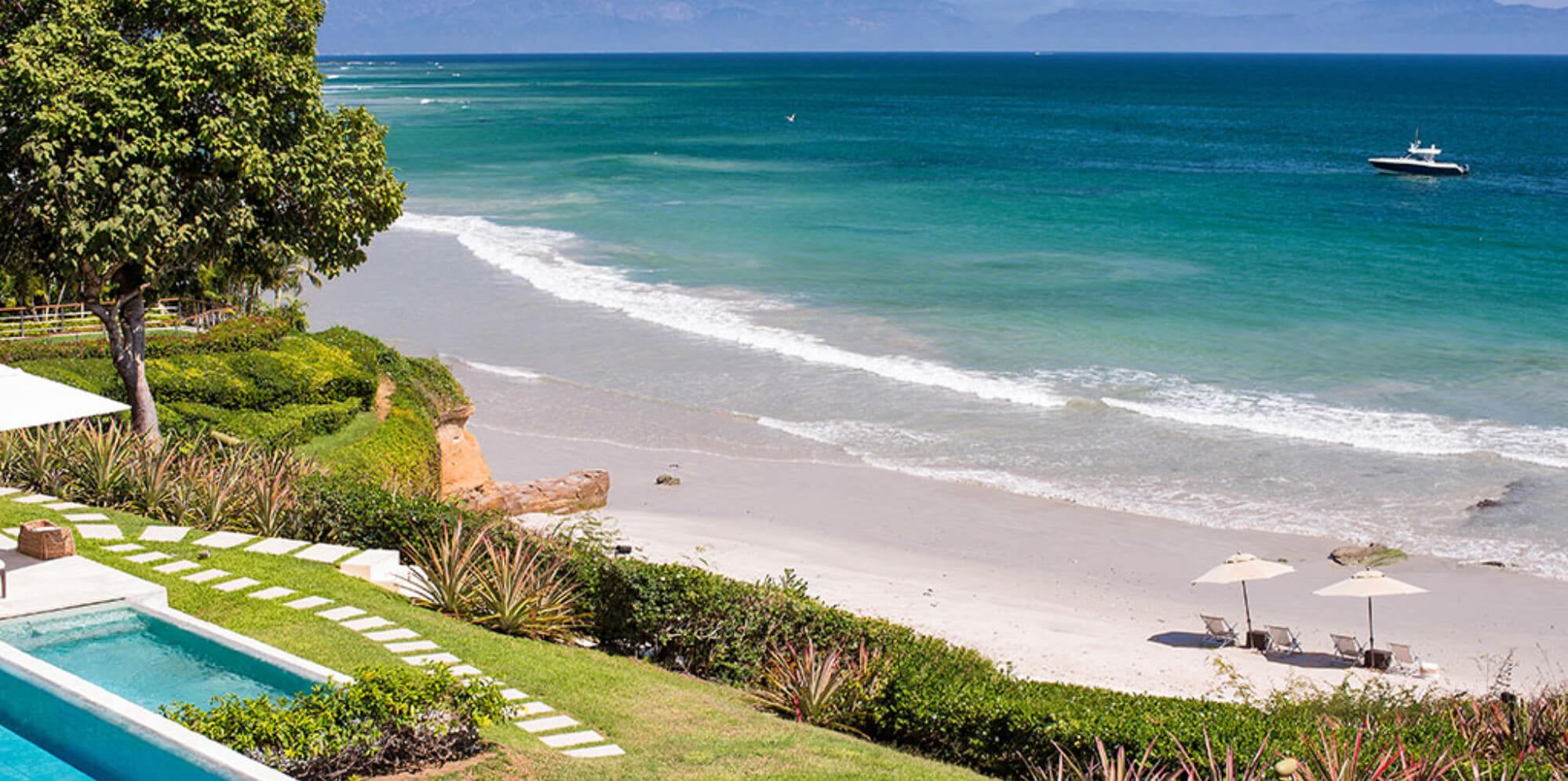 View from the pool terrace over a stone-path garden and agave plantings down to the white-sand private beach and turquoise Banderas Bay with a yacht offshore, Estate Jaguar, Ranchos Estates, Punta Mita.