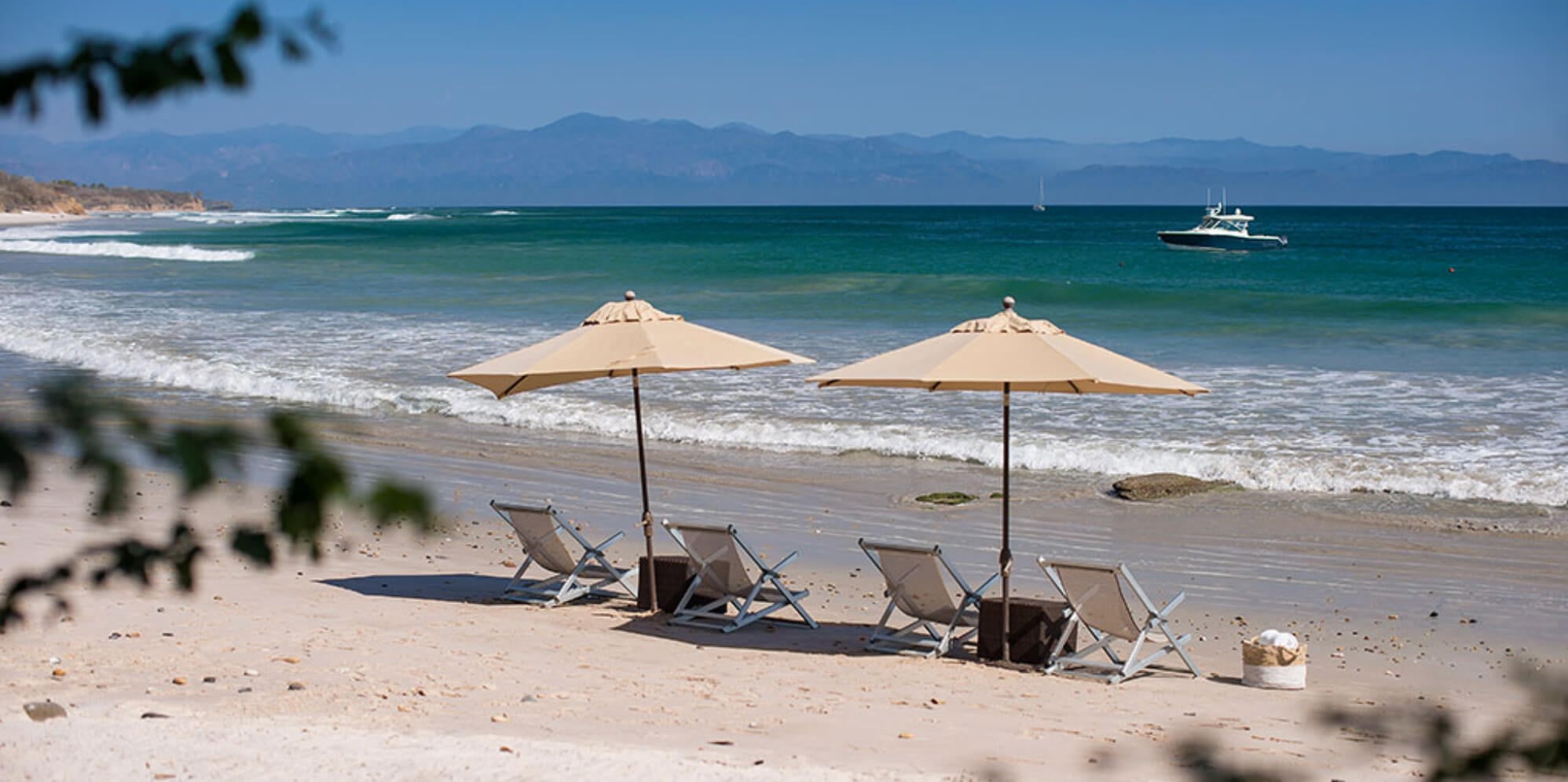 Four beach loungers under two canvas umbrellas on white sand, Pacific surf breaking close, and the Sierra Madre mountains across Banderas Bay, Estate Jaguar, Ranchos Estates, Punta Mita.