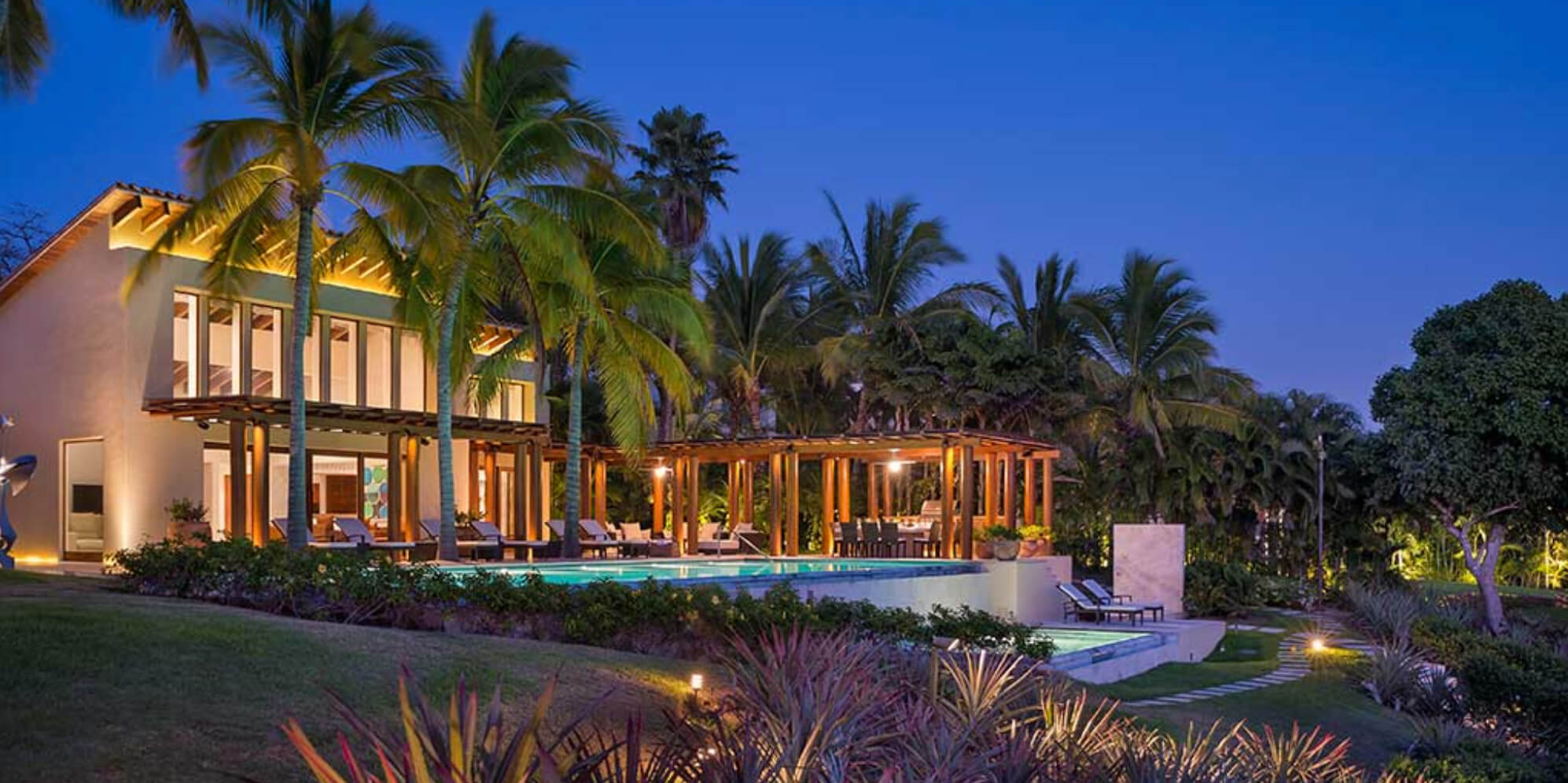 Dusk exterior of cream flat-roof villa with illuminated pool, wood pergola dining structure, and coconut palms under a deep blue sky, Estate Jaguar, Ranchos Estates, Punta Mita.