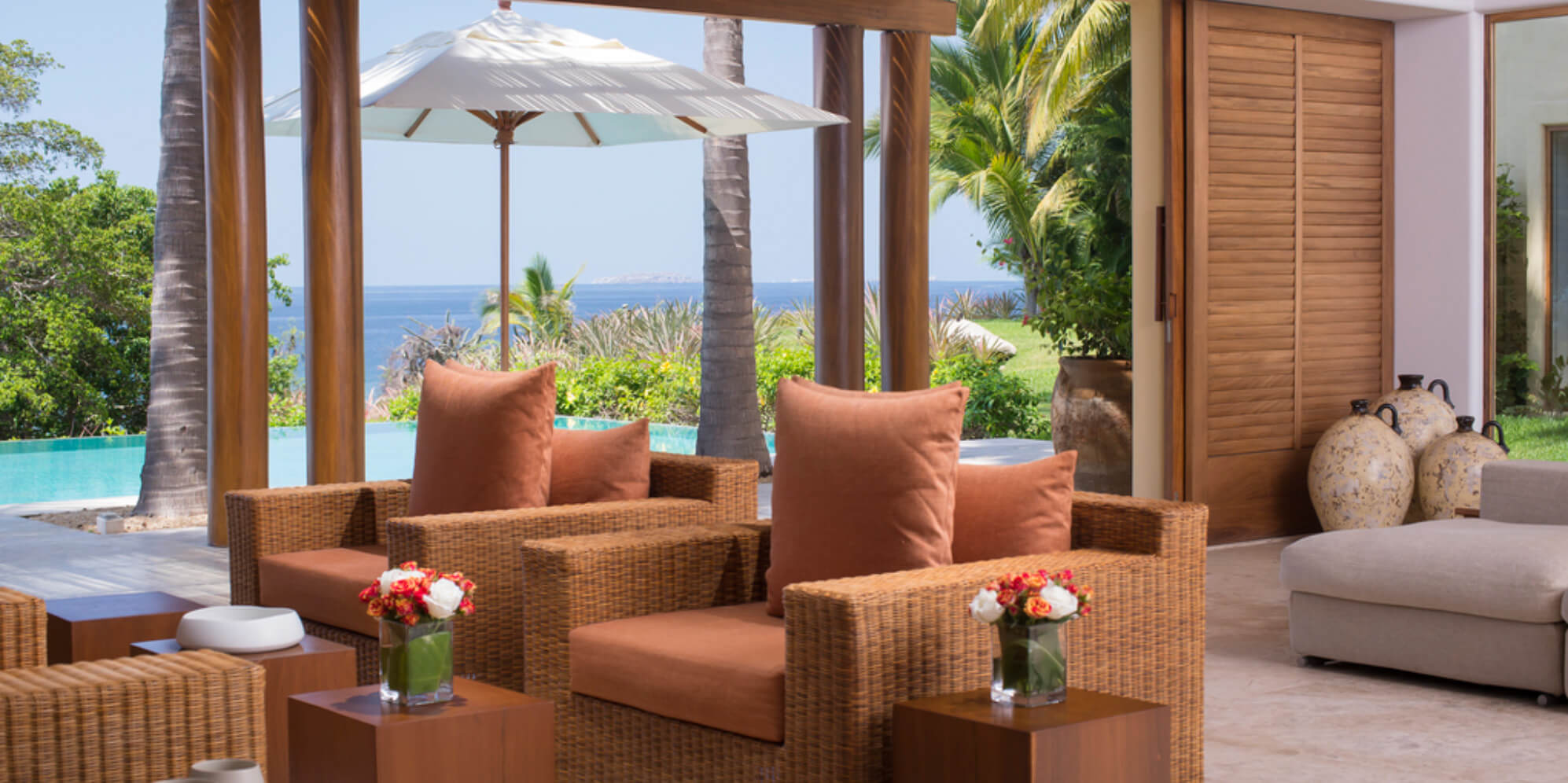 Covered terrace with two wicker armchairs and terracotta cushions under wood columns, pool and Pacific ocean visible beyond, white market umbrella, tropical garden at sides, Estate Jaguar, Ranchos Estates, Punta Mita.