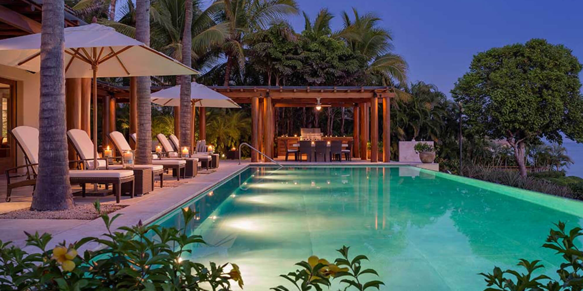 Illuminated rectangular pool at dusk with row of sun loungers under white umbrellas at left and lit wood pergola dining area in the background, Estate Jaguar, Ranchos Estates, Punta Mita.