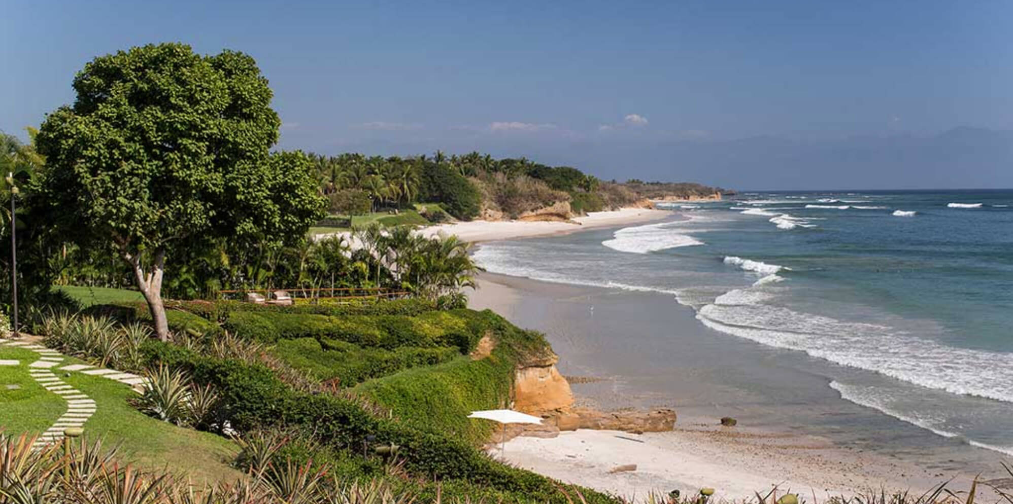 Elevated view from the bluff garden showing a stone-step pathway, manicured agave and lawn plantings, and a wide Pacific beach curving toward distant headlands, Estate Jaguar, Ranchos Estates, Punta Mita.