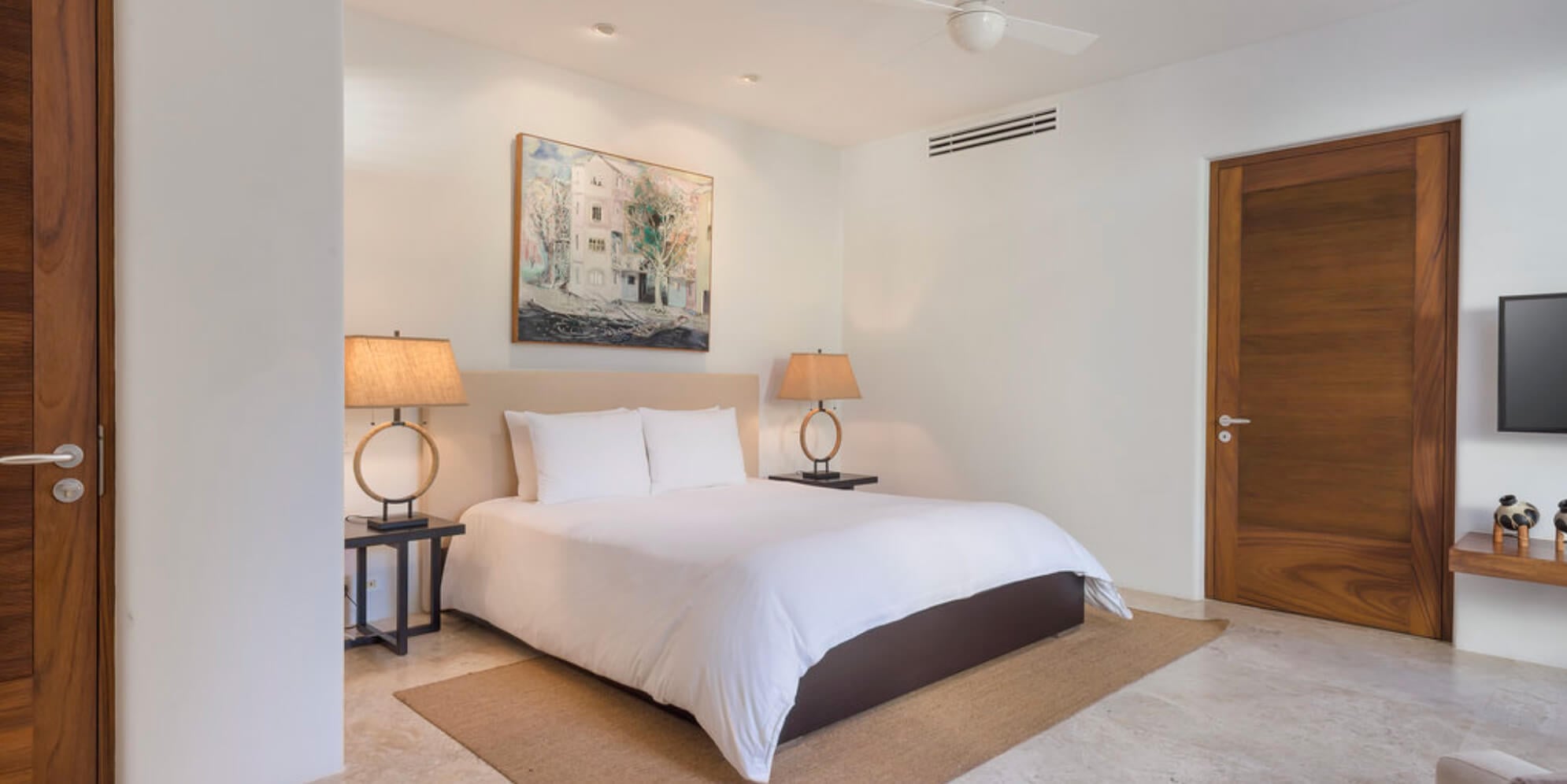 Guest bedroom with cream upholstered headboard, white bedding, paired ring-base table lamps, dark walnut doors, travertine floor, and a figurative painting above the bed, Estate Jaguar, Ranchos Estates, Punta Mita.