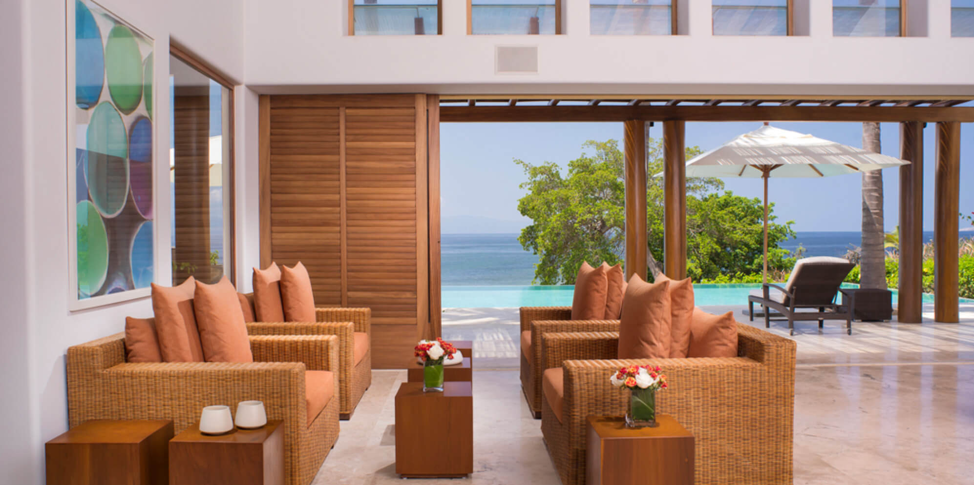Open living pavilion with facing wicker sofas and terracotta cushions, louvered wood panels open to the infinity pool and Pacific, circular colorful artwork on white wall, Estate Jaguar, Ranchos Estates, Punta Mita.