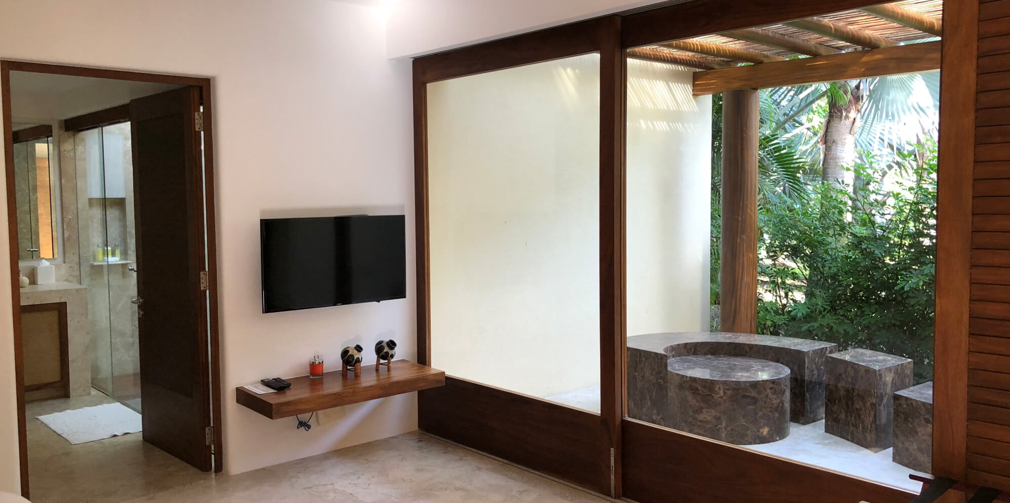 Garden-facing glass enclosure with a dark stone circular soaking tub and matching stool set among palms, wall-mounted television on floating walnut shelf visible inside the bedroom, Estate Jaguar, Ranchos Estates, Punta Mita.