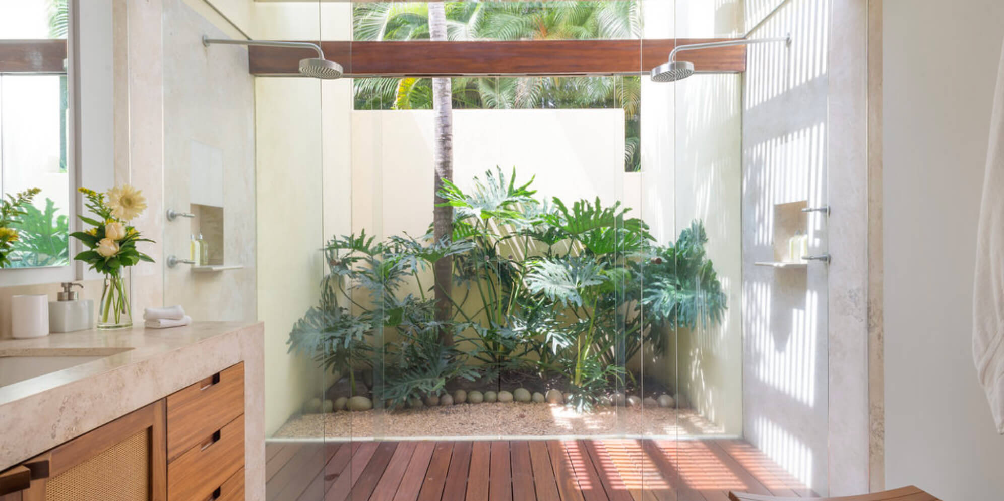 Open-air double rain-head shower with teak deck floor and glass walls enclosing a planted courtyard of monstera and palm, travertine vanity with walnut cabinetry to the left, Estate Jaguar, Ranchos Estates, Punta Mita.