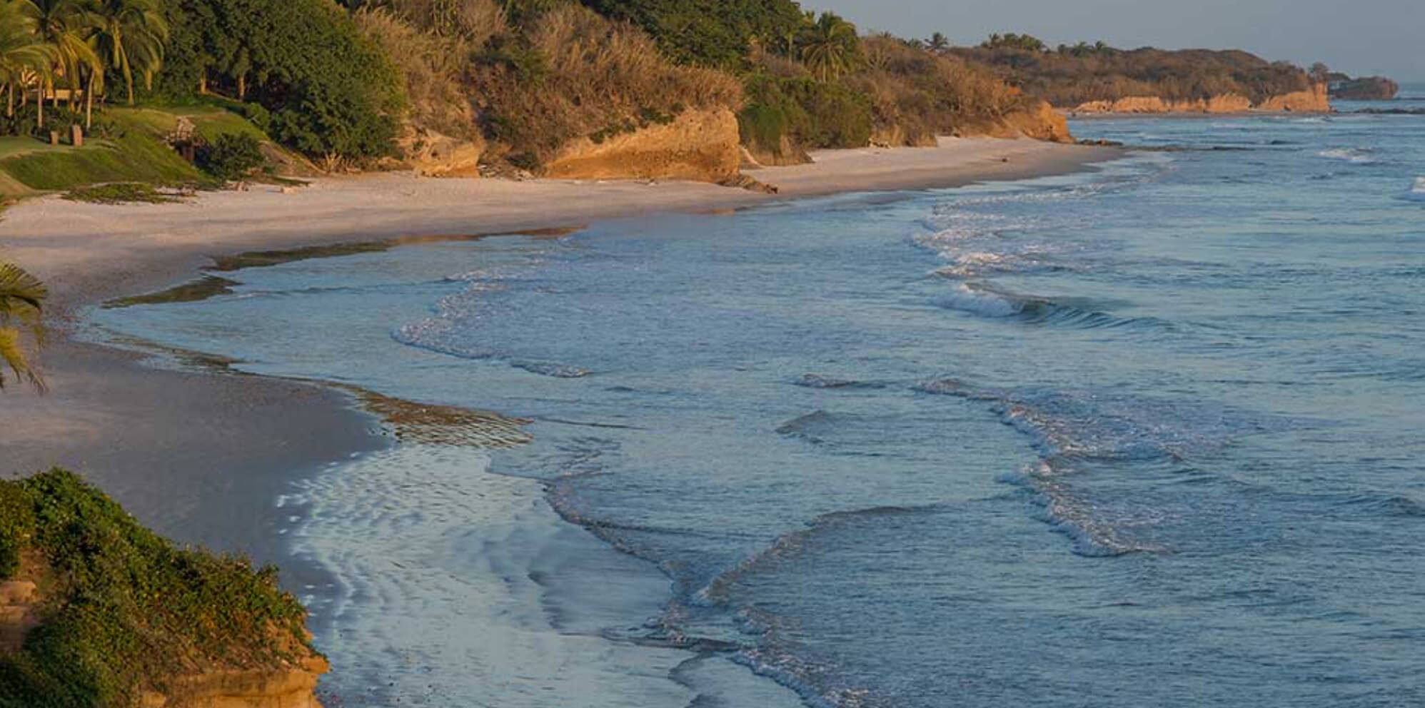 Golden-hour aerial view along the coastline from the property bluff showing white sand beach, Pacific waves, sandstone cliffs, and palm-fringed green grounds at upper left, Estate Jaguar, Ranchos Estates, Punta Mita.