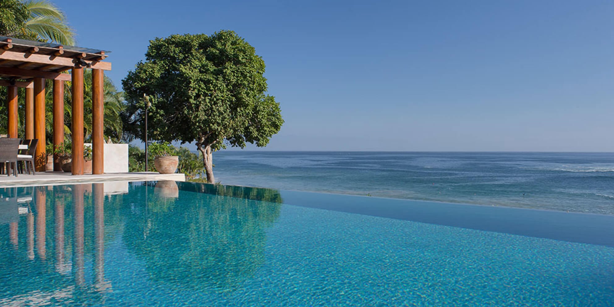 Wide infinity pool surface merging with the Pacific horizon, wood pergola structure and solitary shade tree at the pool edge, sun lounger and terracotta pot visible at poolside, Estate Jaguar, Ranchos Estates, Punta Mita.