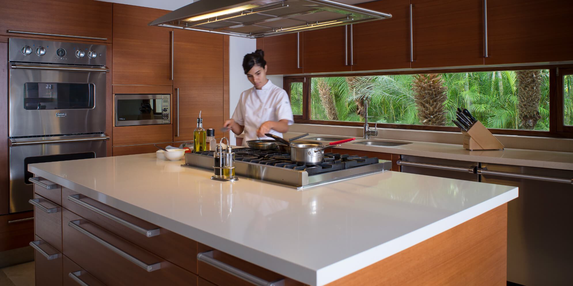 Chef in white jacket working at the gas cooktop island in a walnut kitchen with white quartz surfaces, stainless Viking double oven, ceiling-mounted range hood, and garden-view strip windows, Estate Jaguar, Ranchos Estates, Punta Mita.