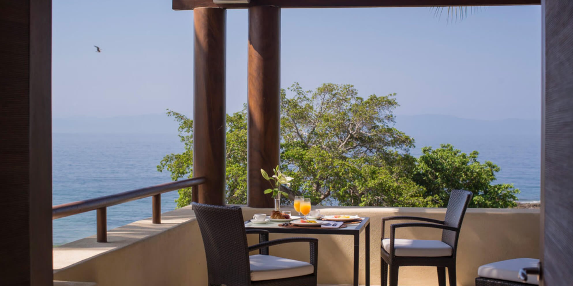 Private suite terrace with two wicker chairs at a bistro table set for breakfast with orange juice and coffee, dark wood pergola framing an ocean view over tropical canopy, Estate Jaguar, Ranchos Estates, Punta Mita.