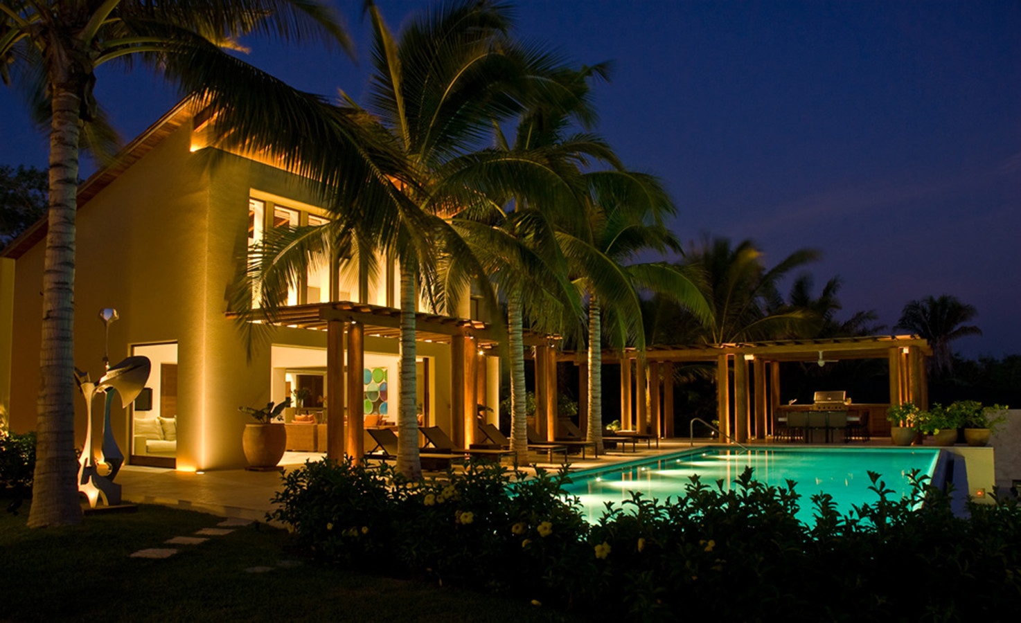 Night exterior showing the illuminated cream flat-roof villa with large pool, wood pergola, mature palms, and a large outdoor sculpture on the side terrace, all glowing under deep blue sky, Estate Jaguar, Ranchos Estates, Punta Mita.