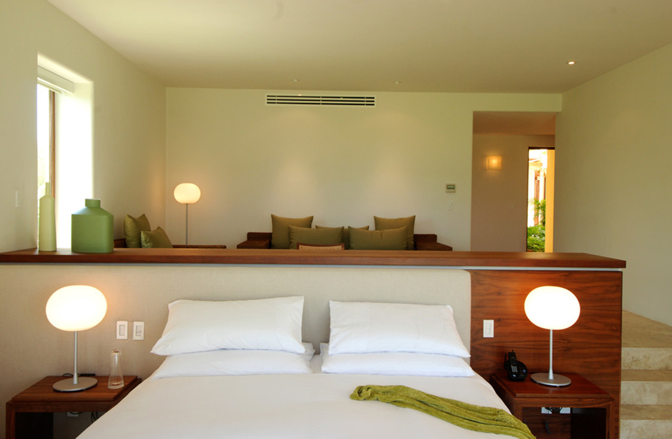 Primary bedroom with built-in walnut headboard and integrated nightstands, globe table lamps, white bedding, and a raised sitting alcove with olive-green cushions behind a walnut partition ledge, Estate Jaguar, Ranchos Estates, Punta Mita.