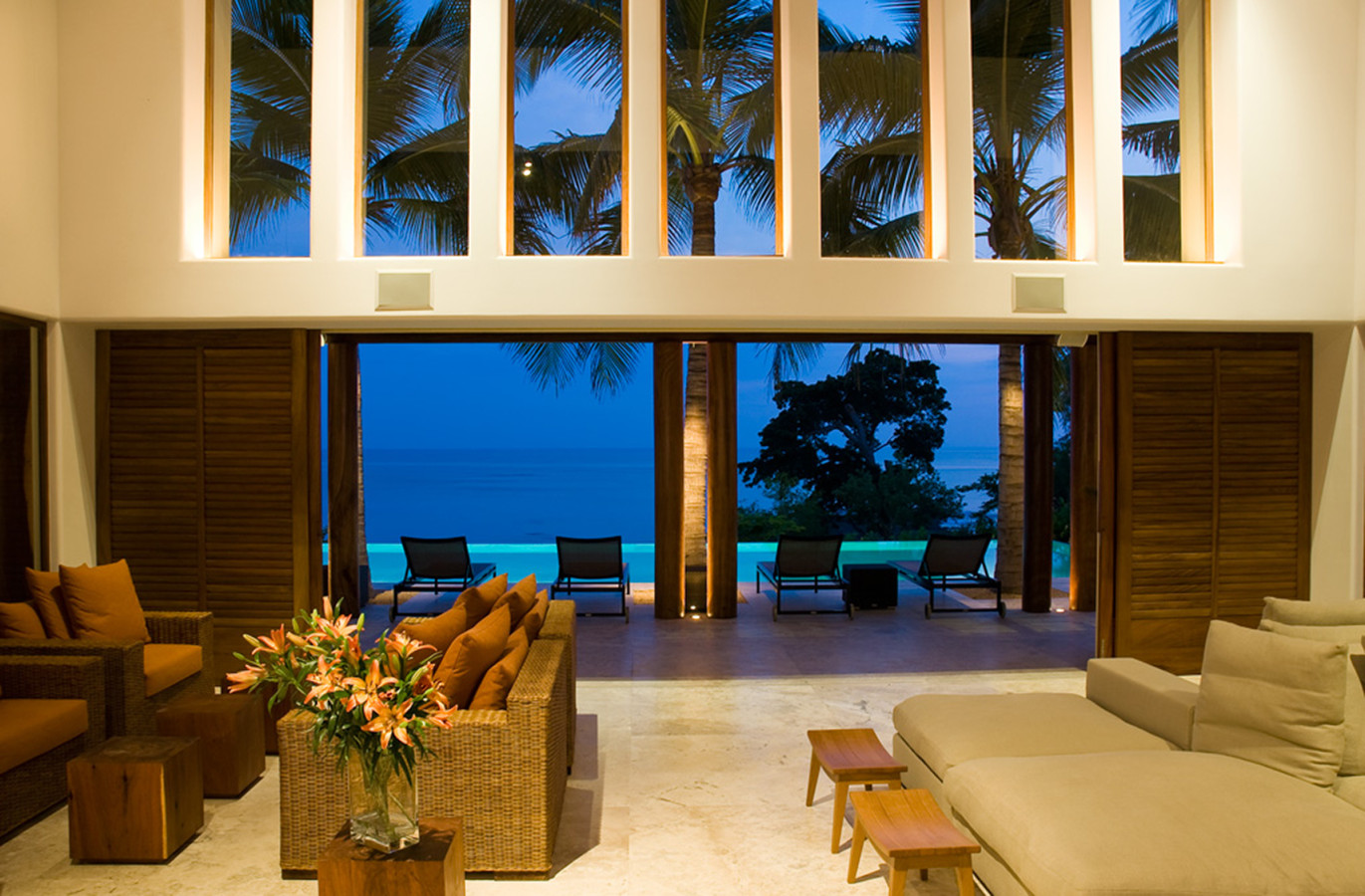 Dusk living pavilion interior with high clerestory windows, wicker seating with rust cushions, orange lily arrangement, and open louvered panels leading to the pool deck and Pacific at blue hour, Estate Jaguar, Ranchos Estates, Punta Mita.