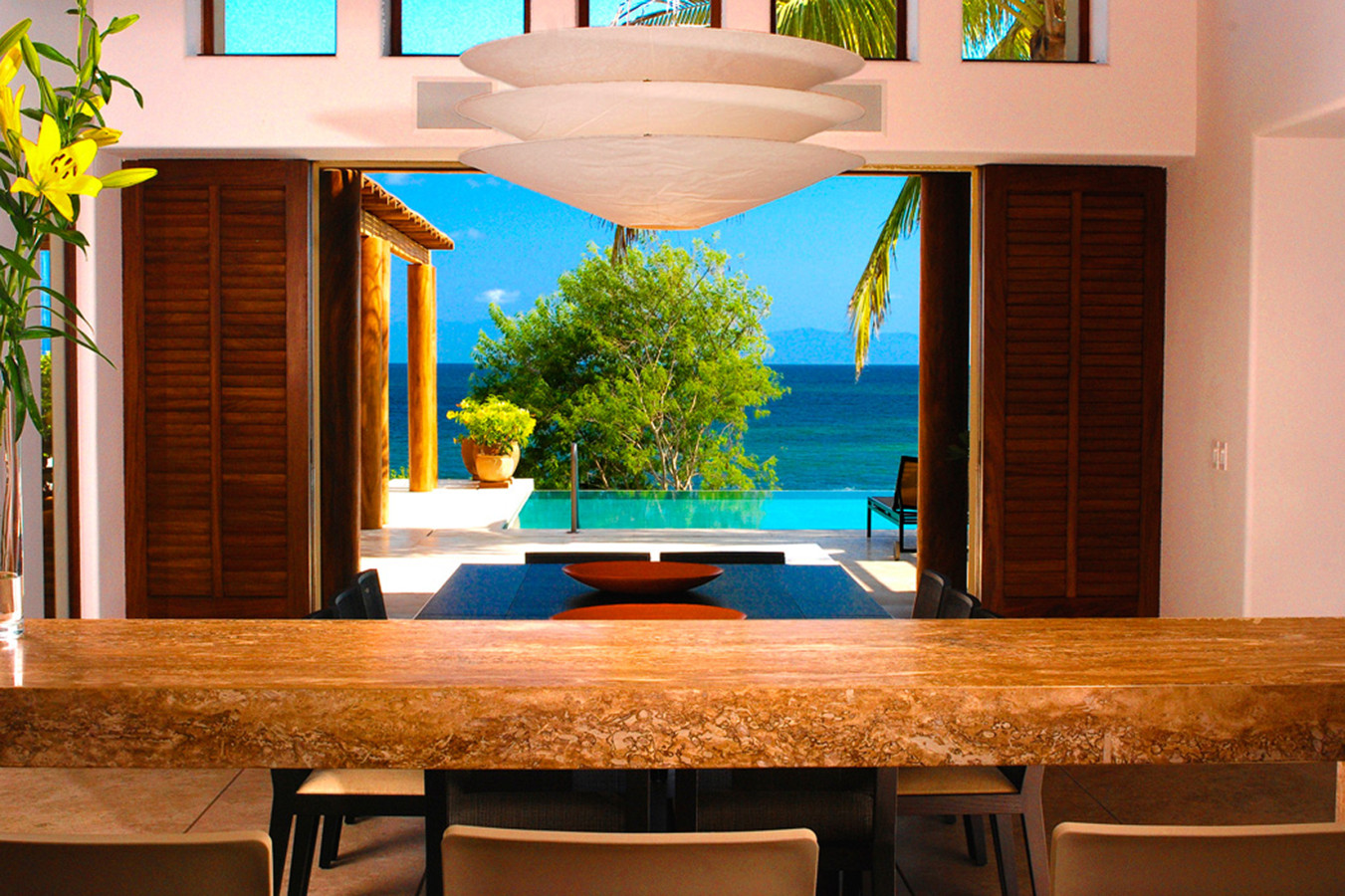 Indoor dining table of travertine stone with dark chairs, large saucer pendant lights, and louvered wood panels fully open to the pool terrace and turquoise Pacific beyond, Estate Jaguar, Ranchos Estates, Punta Mita.