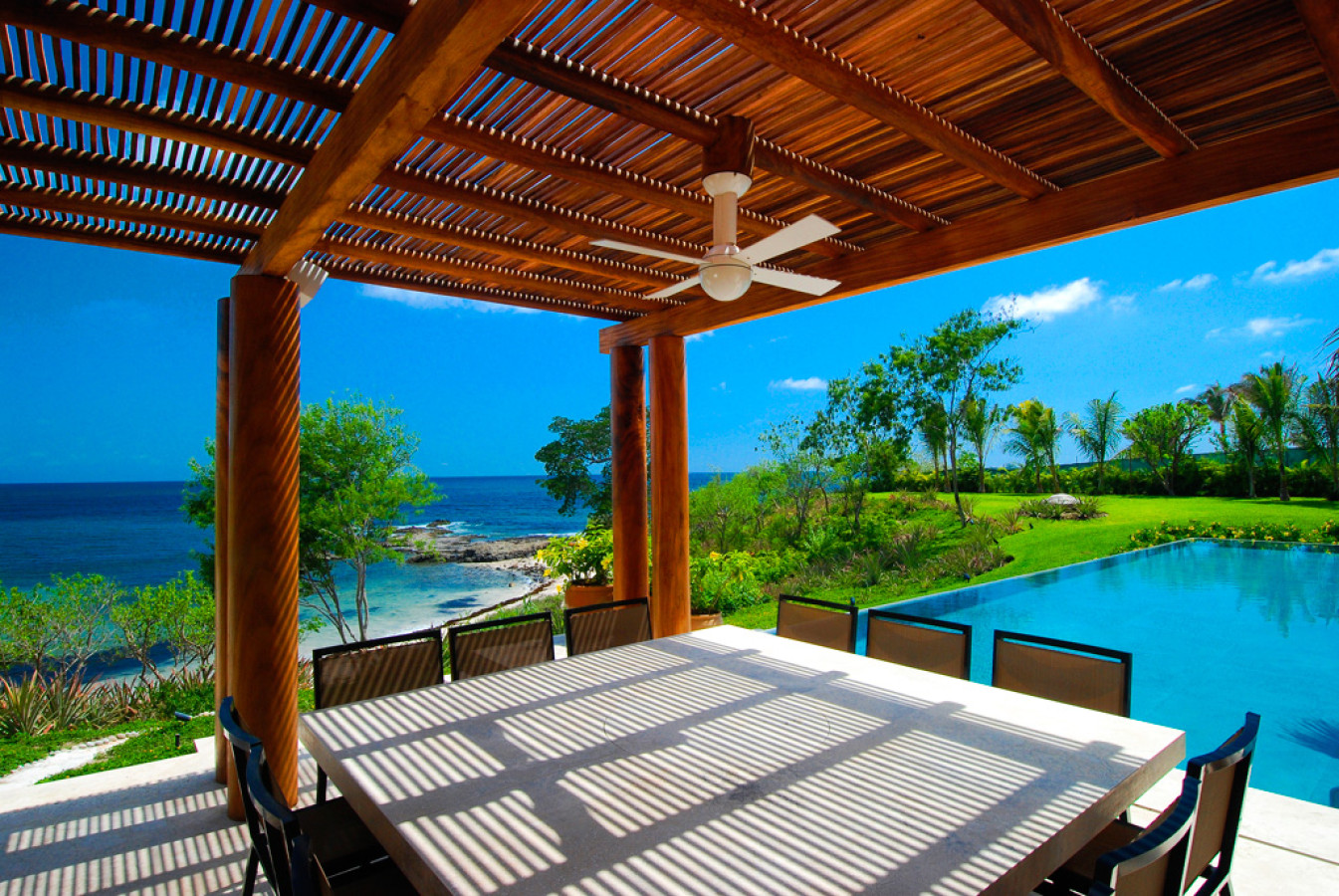 Outdoor dining table under a slatted wood pergola with ceiling fan, eight chairs, pool visible at right, rocky shoreline and Pacific ocean beyond, and tropical lawn in the background, Estate Jaguar, Ranchos Estates, Punta Mita.